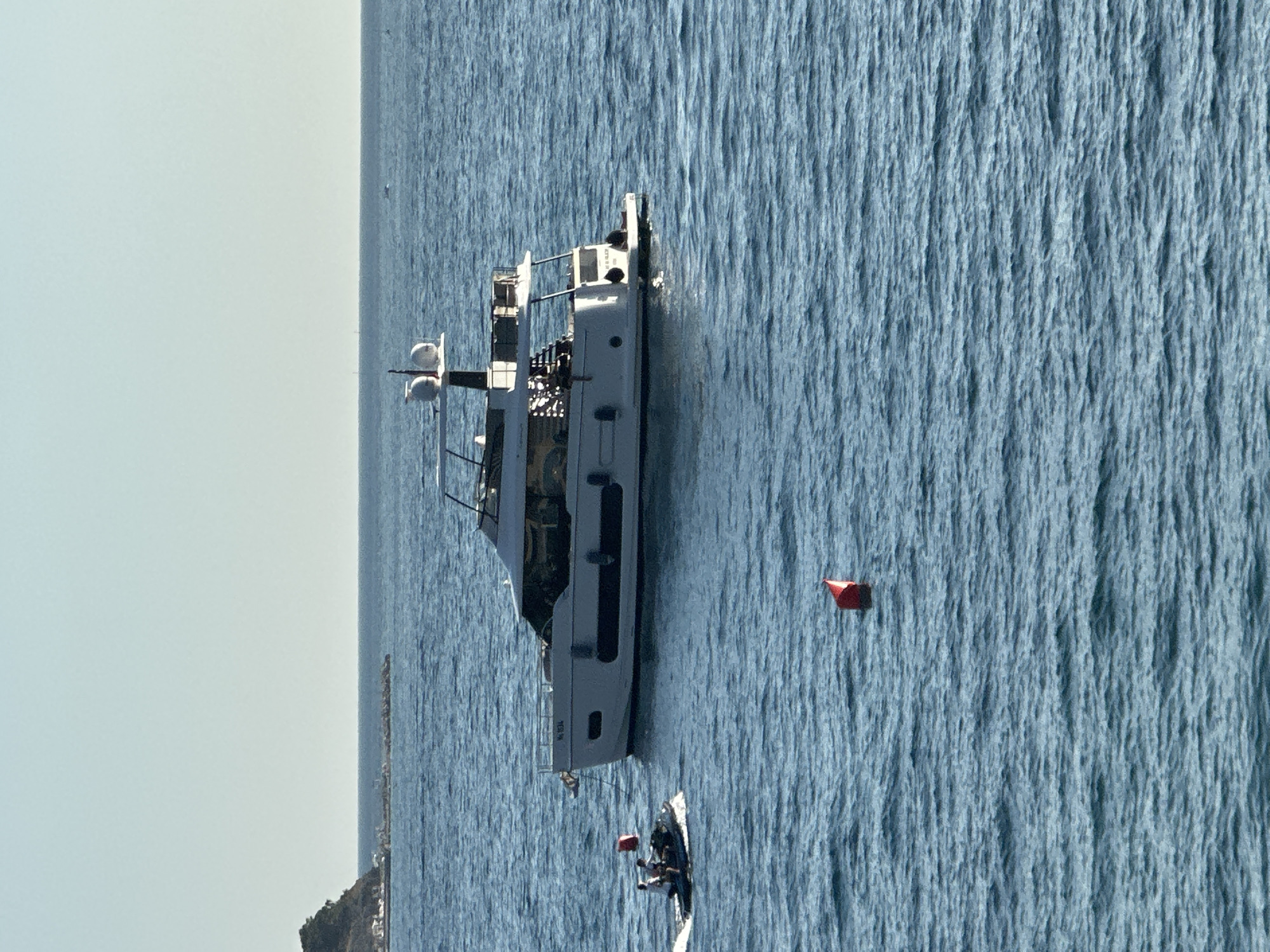 A yacht, identified by the hull number "NL 934," is anchored in the sea, with a jet ski carrying two individuals approaching it. Two red buoys are visible in the water. In the distance, a mountainous, tree-covered island with a small settlement is present. The sky is clear and pale blue. The scene depicts a sunny day at sea, likely near Budva, Montenegro.