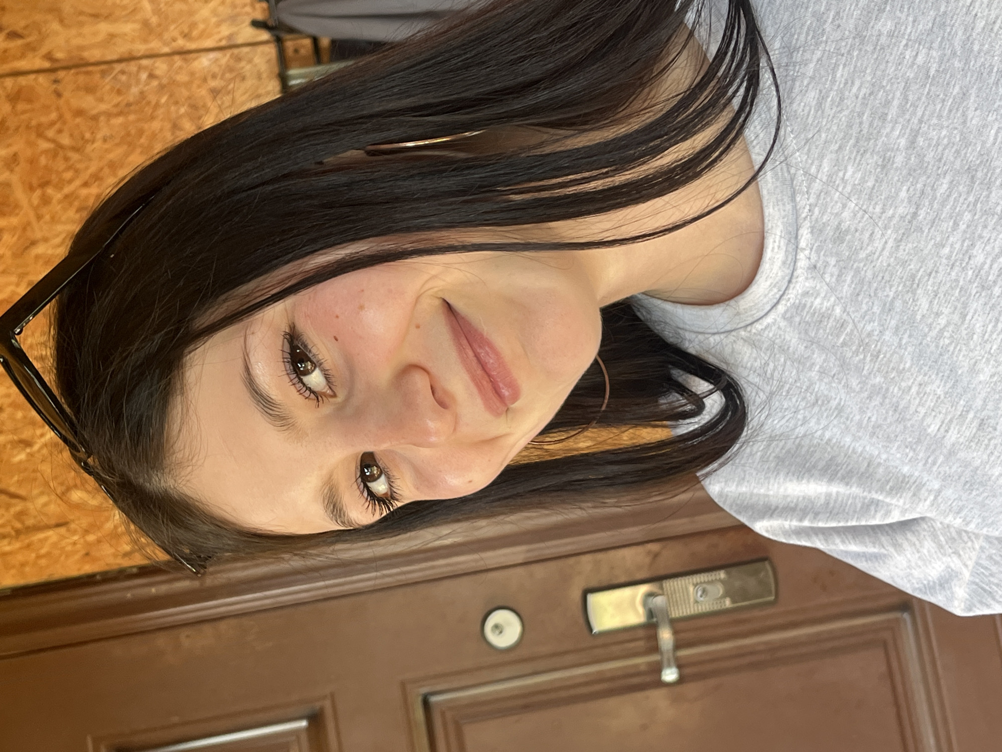 A young woman with dark hair is smiling at the camera. She has brown eyes and fair skin, wearing a light grey T-shirt. Part of a pair of glasses can be seen in her hair. Behind her is a brown door with a golden handle and lock. The background includes wood paneling. Location is unknown and cannot be determined.