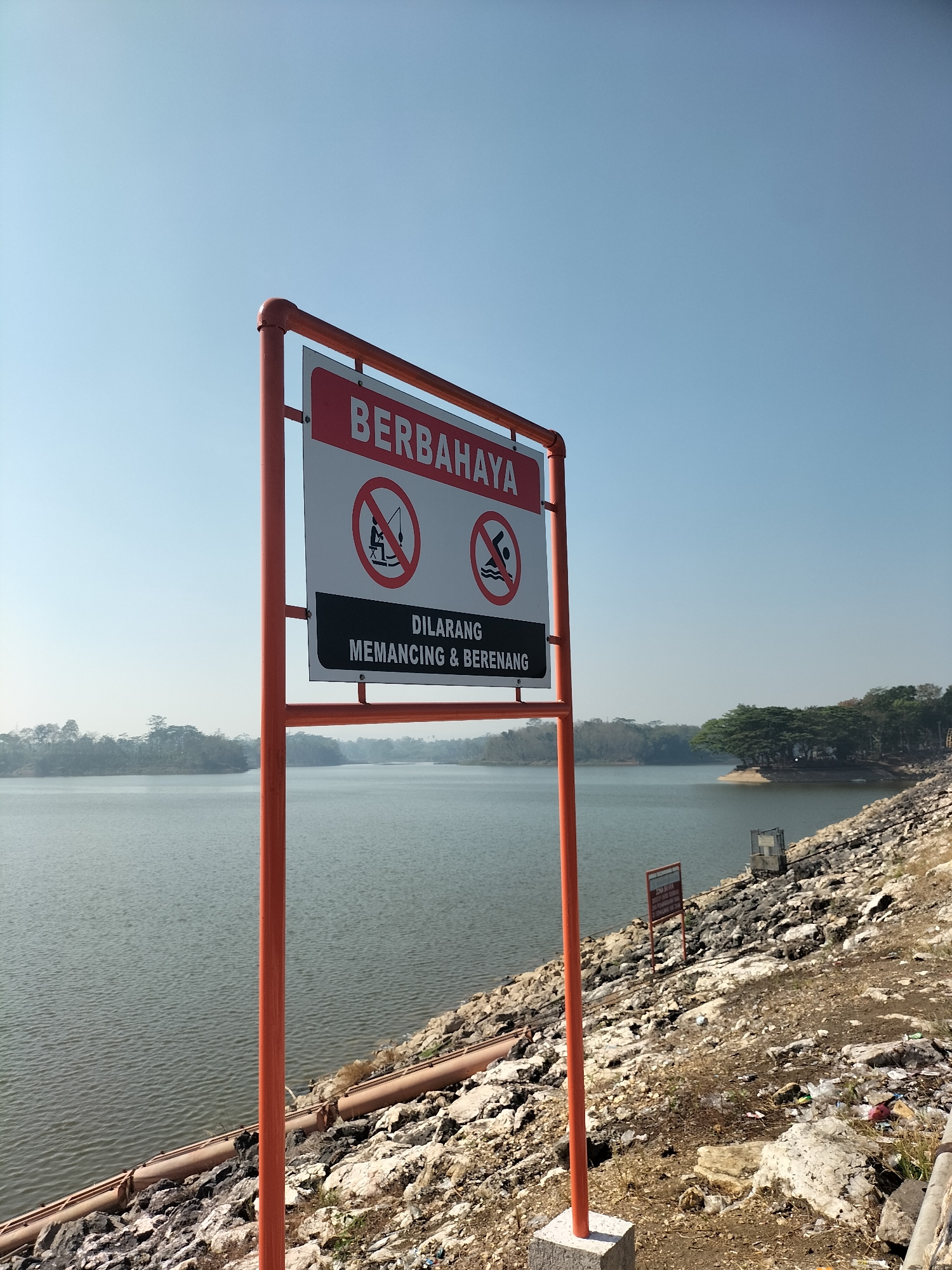A bright, clear day is depicted at a body of water, likely a reservoir, in Sumberpucung, Indonesia. In the foreground, a red metal frame holds a white sign with red lettering that reads "BERBAHAYA" (Dangerous). Below this, two circular prohibition signs are visible: one depicts a fishing rod with a line through it, and the other shows a person diving into water with a line through it. Underneath these symbols, the sign continues in black text with white lettering, stating "DILARANG MEMANCING & BERENANG" (Fishing & Swimming Prohibited). The sign is positioned on a rocky embankment. The water stretches out towards a distant, tree-lined horizon under a clear blue sky. The embankment to the right of the sign is covered in rocks, with some pipes visible along the water's edge and scattered debris. Further in the distance, along the shoreline, some small structures and more trees are present.