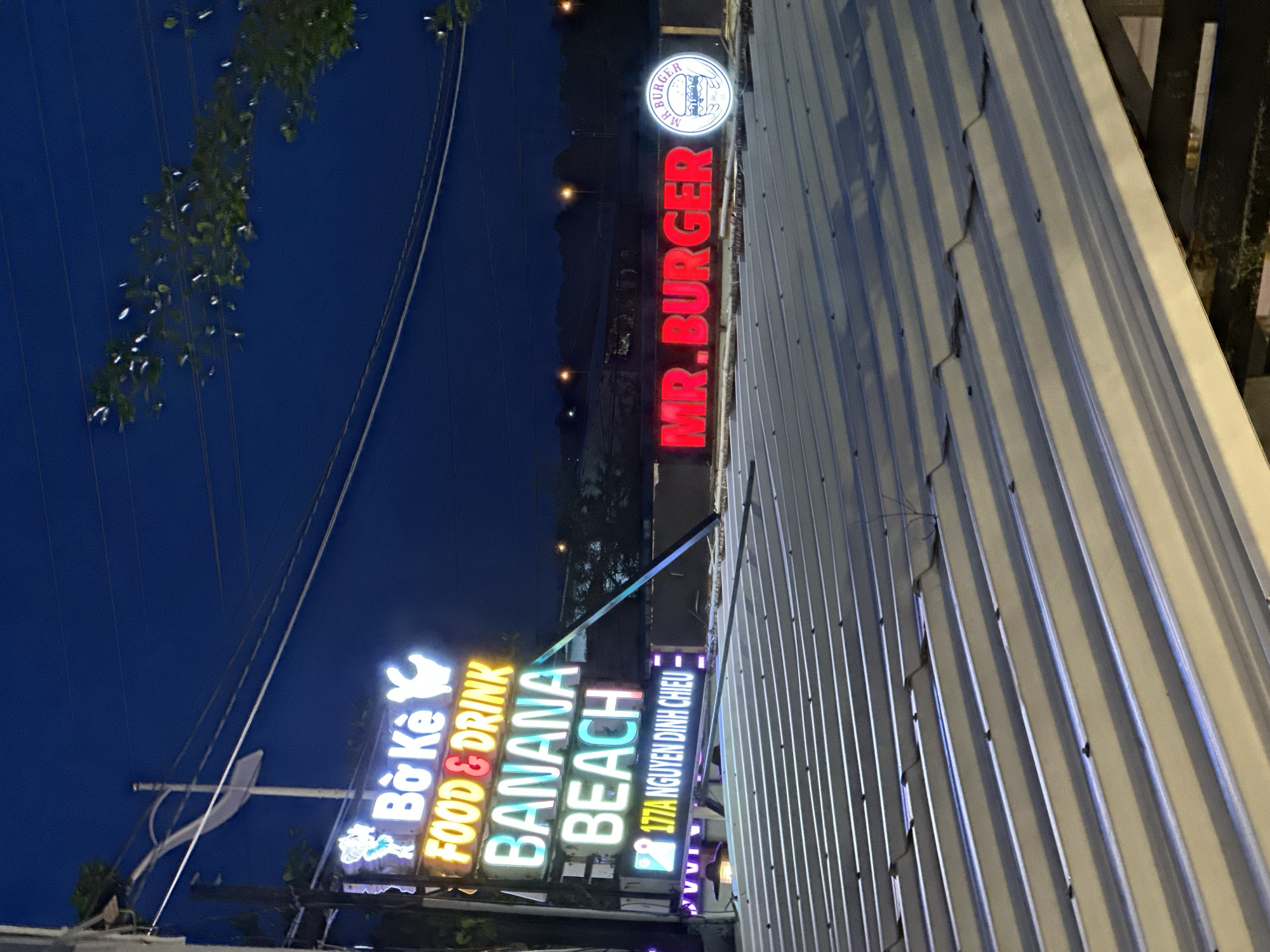 The image shows a nighttime scene in Phu Thuy Ward, Vietnam, featuring illuminated neon signs. To the left, a vertical sign displays "Bo Ke," "FOOD & DRINK," "BANANA BEACH," and "177A NGUYEN DINH CHIEU" in various neon colors. To the right, a horizontal red neon sign reads "MR. BURGER," accompanied by a circular logo depicting a burger. The signs are mounted on poles or a structure that is partially obscured by a corrugated metal roof in the foreground. Overhead, power lines and a street lamp are visible against a dark blue sky. Trees and distant lights suggest an urban or suburban environment.