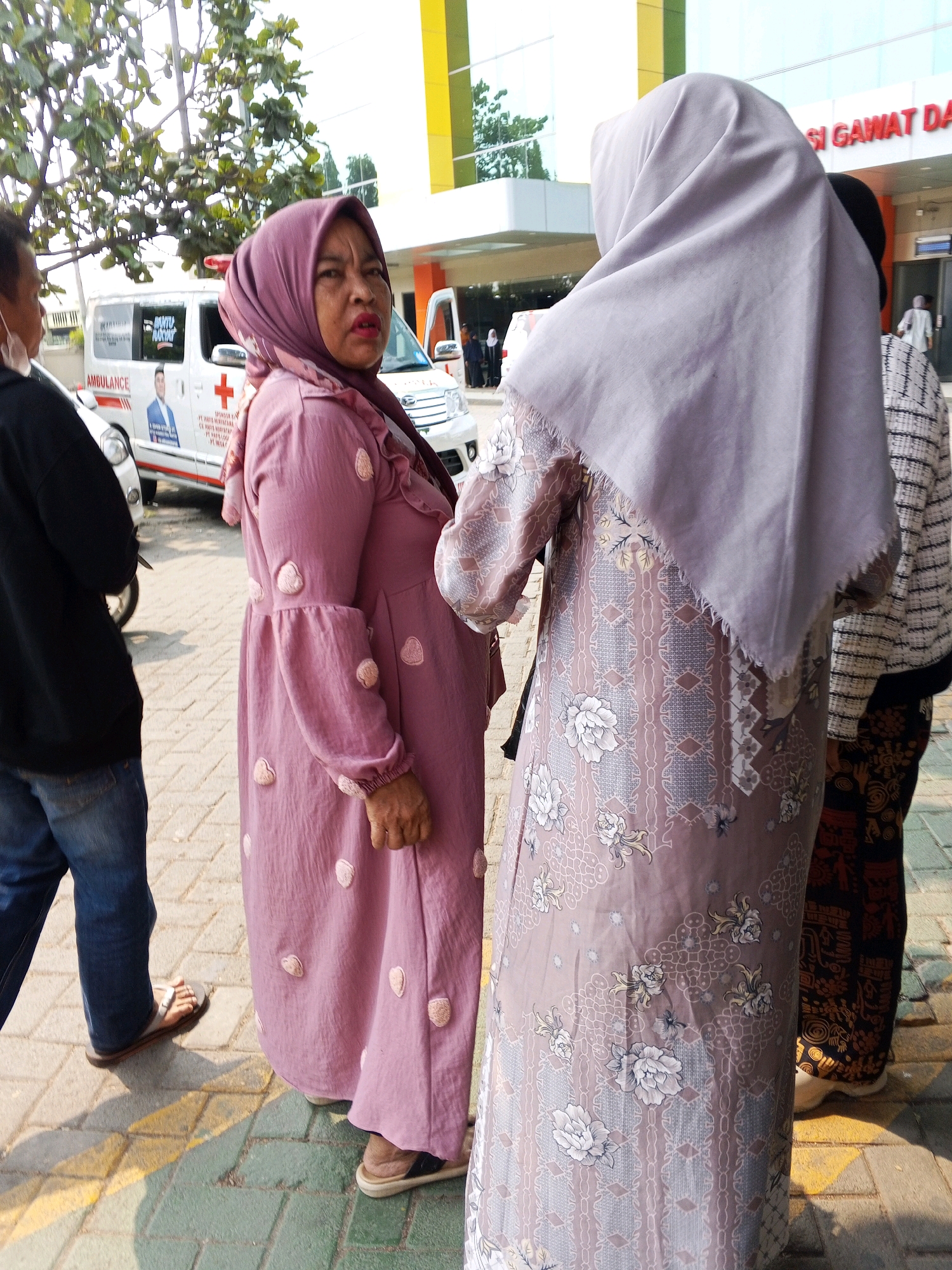 The image shows three individuals standing outside a building with the sign "IGD GAWAT DARURAT" visible, indicating an emergency department. An ambulance with the word "AMBULANCE" on its side is parked to the left. The person in the foreground on the left is wearing a dark long-sleeved shirt and blue jeans, with one foot in a sandal. Beside them, a woman in a purple hijab and a long, flowy purple dress with pink heart-shaped embellishments is looking towards the camera. To the right of her, another person wearing a light purple hijab and a patterned dress stands with their back to the camera, and to their right, a person in a patterned skirt and black and white striped top is partially visible. The ground is paved with interlocking bricks. The building appears to be a hospital or clinic in Ciruas, Indonesia.