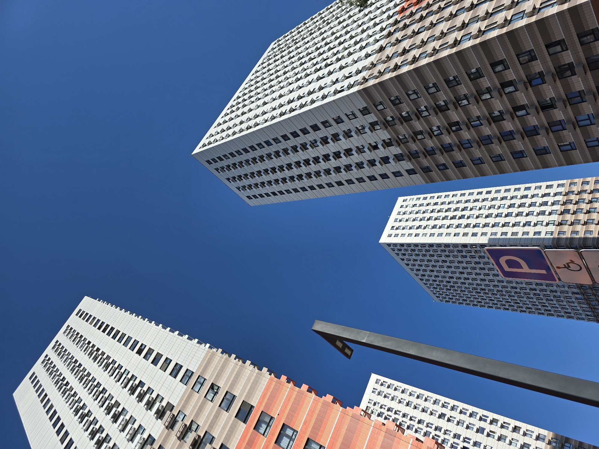 The image shows a low-angle view of several buildings against a clear blue sky. The buildings are modern, with a mix of white, beige, and orange facades. They feature rows of windows. A modern lamppost is visible in the foreground. A sign with a "P" and wheelchair symbol is attached to one of the buildings. The photo was taken in Moscow, Russia.