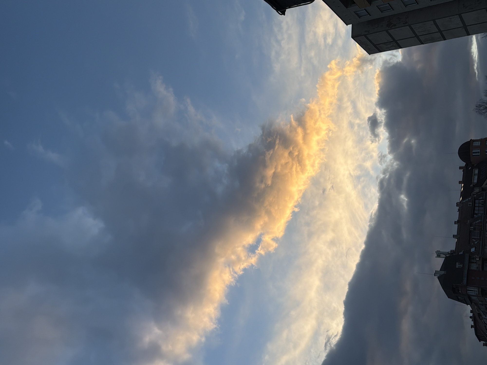The image displays an urban skyscape in Sofia, Bulgaria, under conditions indicative of either sunrise or sunset. The upper two-thirds of the frame are dominated by a cloudy sky. A prominent, elongated cloud formation, brightly illuminated with intense orange and yellow light, stretches diagonally across the mid-section from the upper left to the lower right. This radiant cloud is set against a backdrop of darker, unlit grey and white clouds, with patches of clear light blue sky visible, particularly in the upper right quadrant.
The lower third of the image reveals a mixed architectural environment. On the left, a series of multi-story residential or commercial buildings feature traditional European design, characterized by dark, steeply pitched roofs, visible dormer windows, and several chimneys. One distinct building exhibits a decorative, dark dome-shaped turret at its visible corner. Several slender antennae or lightning rods protrude from the rooftops. In stark contrast, a segment of a tall, grey, unadorned concrete apartment building, likely a residential block with rectilinear windows, is visible on the far right side of the frame. No individuals, specific actions, or interactions are discernible. A few bare tree branches are visible at the bottom edge, suggesting a cooler season.