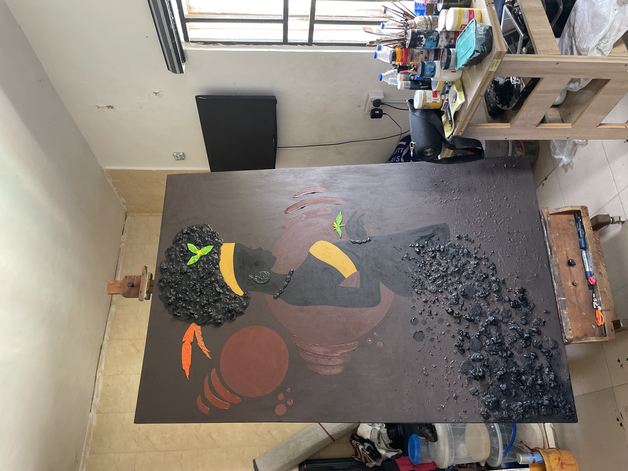 The image shows a painting in a studio in Ado, Nigeria. The painting depicts a woman lying on a dark brown surface, with a hairstyle made of black textured material. She has a yellow armband and a dark necklace. A stylized green leaf sits on her hand, and a larger, brown circular shape with curved lines is below her. Orange and brown crescent shapes are placed on the lower left. The top right corner of the painting has a rough texture made of what looks like pebbles, creating a textural contrast. The painting is on an easel.
The studio environment includes an art supply cart filled with various paints and brushes, a black television on the wall, a window with bars, and assorted containers and materials scattered on the floor. The floor is tiled.