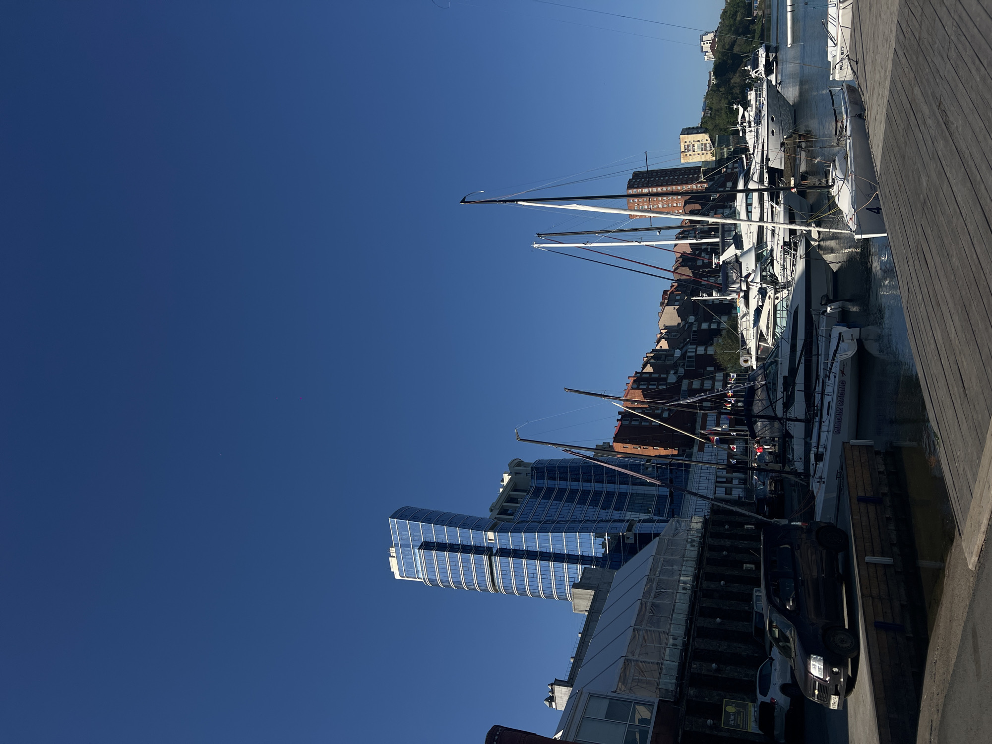 The image depicts a waterfront scene in Vladivostok, Russia, characterized by docked yachts and urban architecture. A modern, glass-clad skyscraper stands prominently in the lower portion of the image, contrasting with older, brick buildings further along the harbor. Numerous sailboats and yachts are docked along a wooden pier that runs along the right-hand side of the frame. A dark-colored vehicle is parked near the edge of the dock. The sky above is a clear, deep blue.