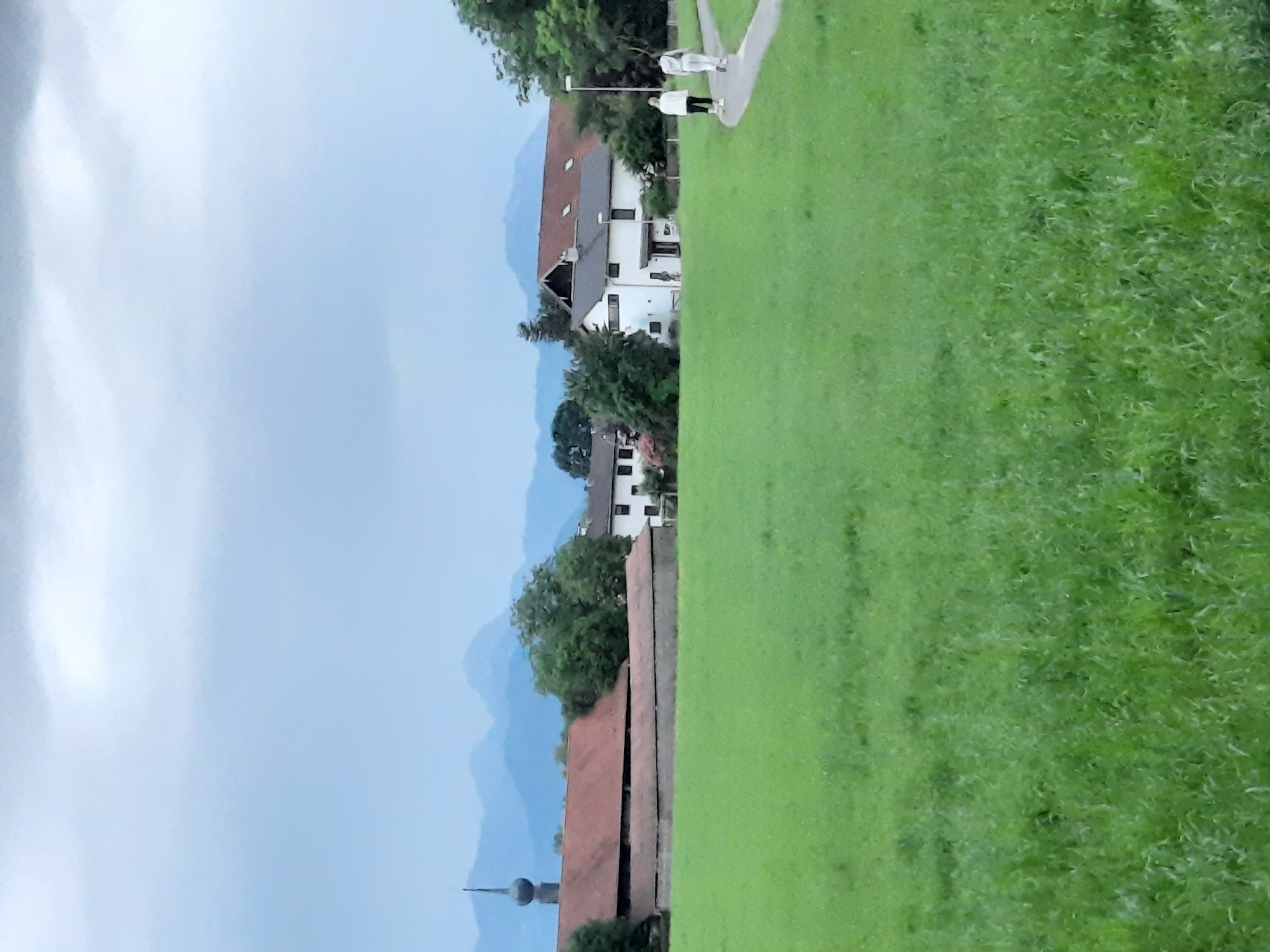 An outdoor, full shot, medium-low angle, depicts a pastoral scene with a verdant green meadow occupying the foreground. A winding paved path cuts through the meadow, and two figures, walking away from the camera, are visible on the path. The figures are dressed in casual, light-colored clothing. In the middle ground, various buildings are present, including a long, barn-like structure with a red roof and a tall, slender spire, possibly a church tower, to its left. Adjacent to the barn are white residential buildings with pitched roofs. Trees are interspersed among the buildings. The background features a hazy, layered view of mountains under an overcast, grey sky. The overall impression is one of a rural or semi-rural setting, possibly on the outskirts of a town. This scene appears to be located in the vicinity of Rosenheim, Germany, given the context of the query.