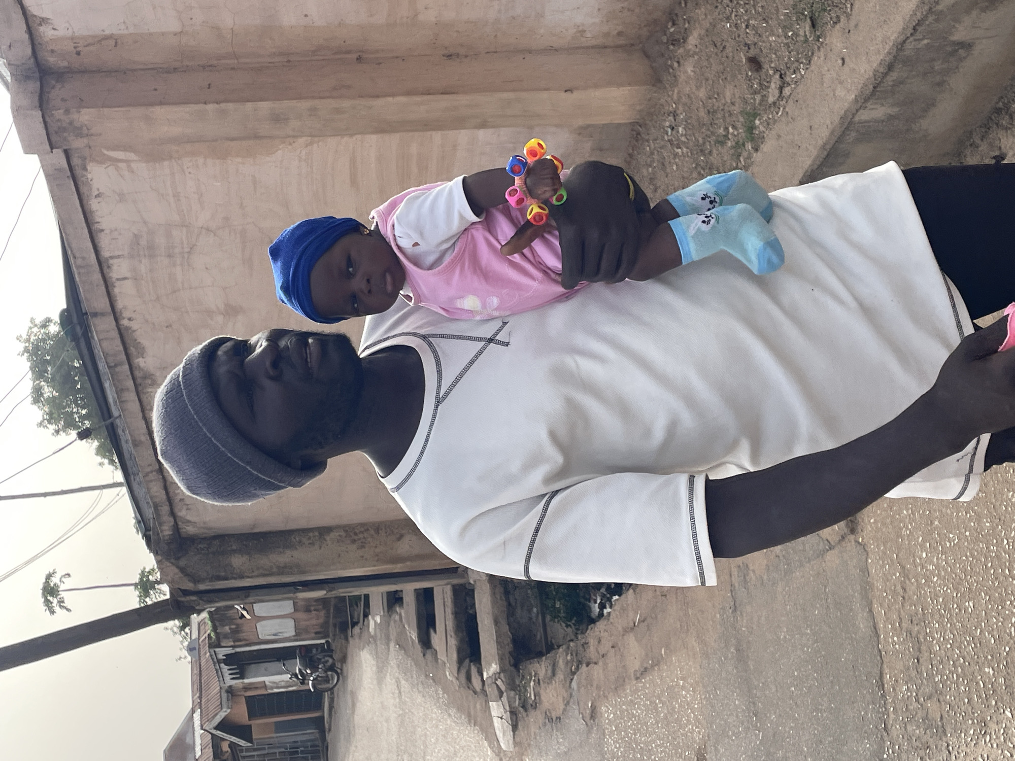 In Jos, Nigeria, a man wearing a gray beanie, white t-shirt, and black pants holds a baby in his arms. The baby is wearing a blue cap, pink dress, and light blue socks. The baby is holding a colorful toy in their hand. They are on a street lined with buildings.