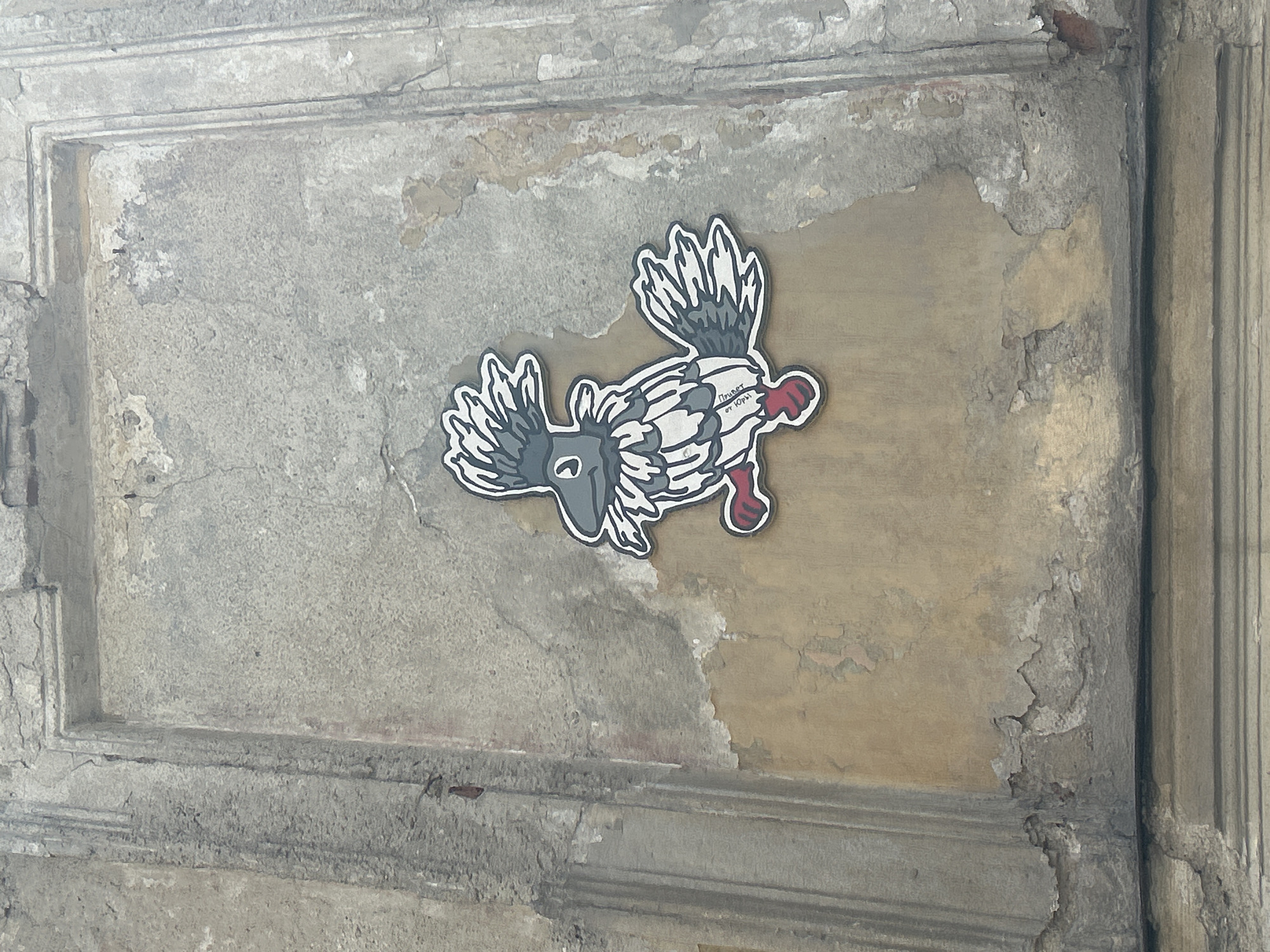 The image shows a cartoon bird sticker adhered to a weathered, textured wall. The wall is composed of large, roughly plastered rectangular sections, with visible signs of age and damage, including cracks, peeling paint, and areas where the plaster has fallen away, revealing brick underneath. The bird sticker is depicted in a cartoonish style, with a gray head and body, white and gray patterned feathers, red feet, and a crest of gray feathers. The bird is facing to the left and appears to be in a walking or hopping pose. The overall impression of the scene is one of urban decay and street art. The image is taken from a slightly upward angle, with the top of the wall visible at the top of the frame.
While the prompt requested location information for Perm, Russia, this image itself does not contain any visual cues or text that would indicate its location within Perm. Therefore, it is not possible to determine the specific location in Perm from the provided image.