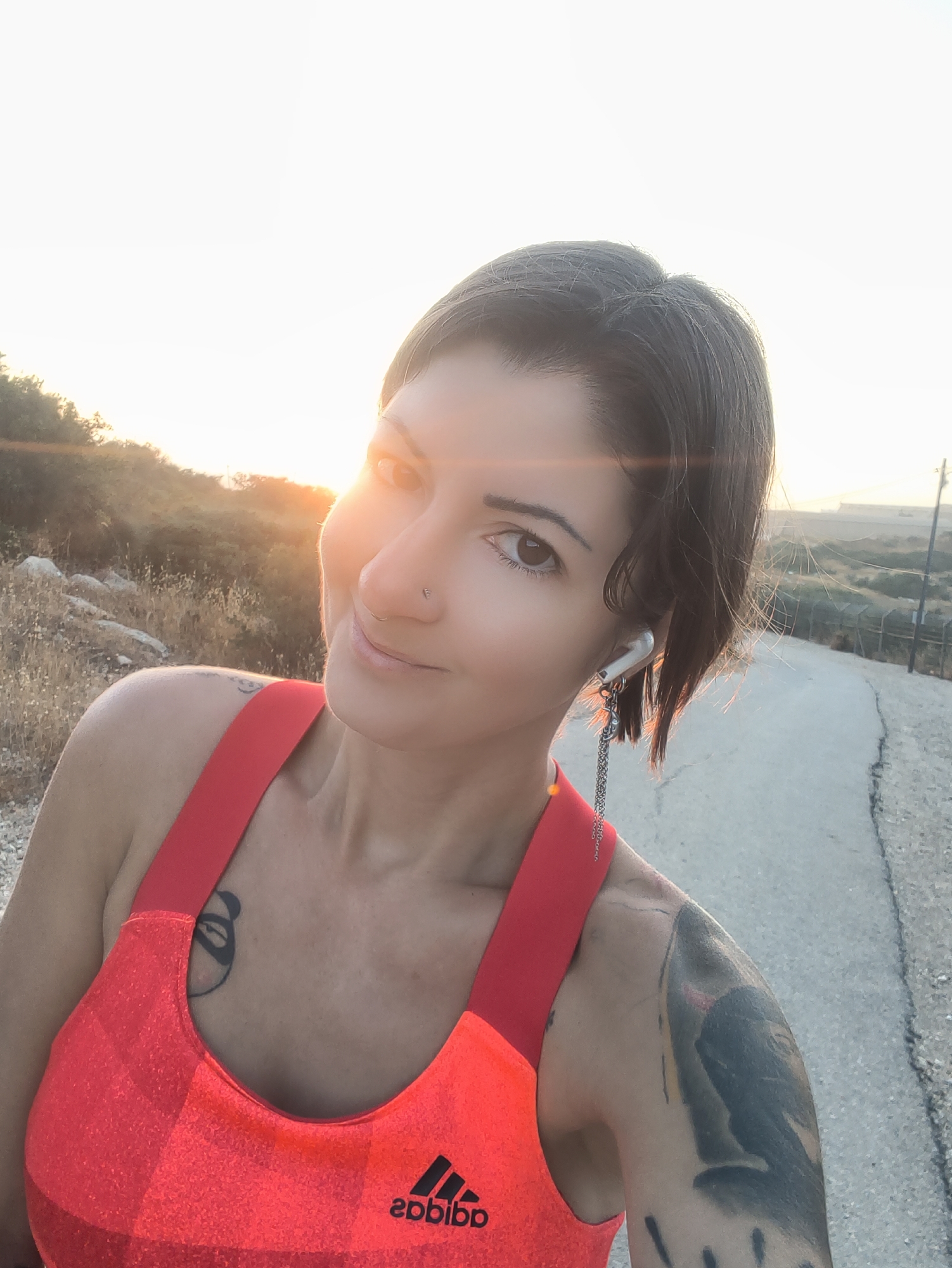 A woman is taking a selfie outdoors on a road. She is wearing a bright orange athletic top and has tattoos on her chest and arm. She is also wearing an earbud with a decorative chain attached. The background shows dry, hilly terrain with sparse vegetation, and a paved road stretching into the distance. The sun is setting or rising, casting a warm glow and lens flare across the scene. The location is identified as the Lachish Regional Council, Israel.
