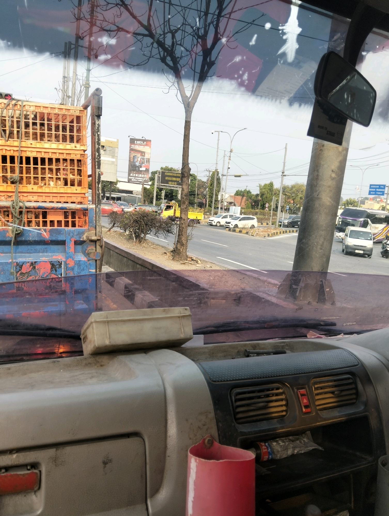 This is a photograph taken from the interior of a vehicle, looking forward through the windshield. The scene is a street in Semarang, Indonesia. In the foreground, on the dashboard, is a small, weathered, light-colored plastic container. The dashboard itself is a dull gray material, with visible dust and wear. To the right of the container are air vents and a hazard light button. A red cup or container is visible on the lower right of the dashboard, and a tube of what appears to be toothpaste or sealant is in a compartment below the air vents.
Through the windshield, we see a truck on the left, loaded with stacked orange crates containing live poultry, likely chickens. The truck's blue bed is visible, showing signs of rust and wear. Beyond the truck, a road is visible with various vehicles including cars, buses, and motorcycles. A yellow truck is driving on the road. Traffic lights and street lamps are present. In the background, there are buildings and trees, indicating an urban environment. A billboard with Chinese characters and an image of machinery is visible on the right side of the street. The sky is overcast. Reflections are visible on the windshield, including the silhouette of tree branches and some abstract shapes.