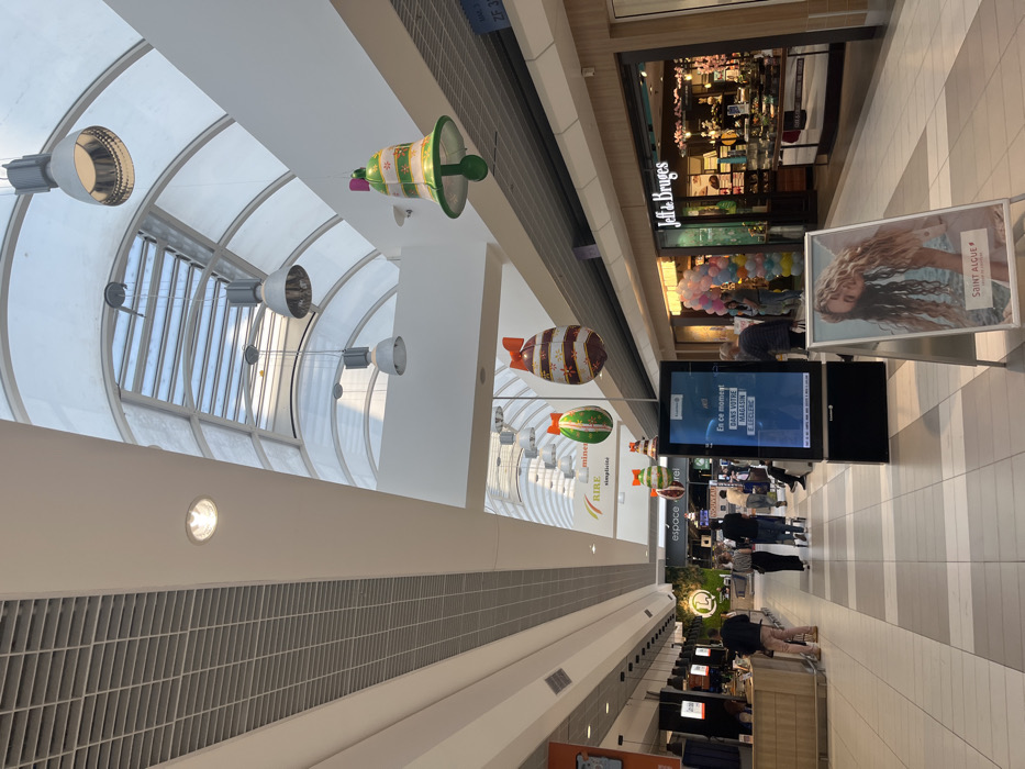 The image shows the interior of a shopping mall in Saint-Brice-Courcelles, France, located at coordinates 49.283, 4. The mall has a vaulted ceiling with a large skylight. There are multiple hanging light fixtures and festive decorations, including bells and Easter eggs. Several people are walking through the mall, passing various stores and kiosks. There is a large digital display on the left side of the image displaying a message in French. In the foreground, there is an A-frame sign advertising a brand called Saint Algue. On the right side of the image, there is a store called Jeff & Bruges.