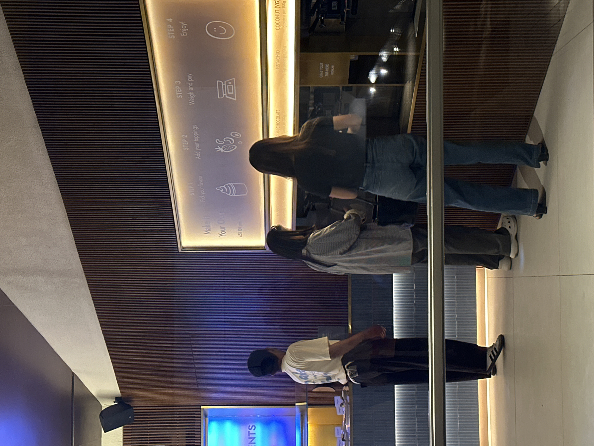 The image depicts an indoor scene, likely a self-serve dessert or frozen yogurt shop, with three people standing in line, facing a counter. The setting appears modern, characterized by dark, horizontally slatted wall paneling and a lighter concrete or stone-like ceiling. Strip lighting runs along the top of the counter and illuminates various signs.
The first person in line, closest to the viewer, wears a light-colored t-shirt, dark trousers, and a dark cap, with black and white striped sneakers. The second person has long dark hair, wearing a striped long-sleeved top over a lighter garment, dark trousers, and white slip-on shoes. The third person, further down the line, also has long dark hair, wearing a dark top, blue jeans, and dark shoes. All three individuals have their backs to the camera, appearing to be engaged in the process of ordering or preparing their dessert.
Above the counter, a prominent illuminated sign outlines a four-step process: "STEP 1 Pick your flavour" (with an ice cream cone icon), "STEP 2 Add your toppings" (with icons of fruit and candy), "STEP 3 Weigh and pay" (with a weighing scale icon), and "STEP 4 Enjoy!" (with a smiley face icon). Below these steps, flavor options are listed, including "GIAN CHOCOLATE," "PISTACHIO," and "COCONUT (VG) 1g protein per 100g." Another sign says "GRAB YOUR TUB HERE REGULAR." A partial sign on the left reads "ENTS," and another sign indicates "Make Your Own" with a price of "45K IDR," suggesting the location is in Indonesia. The artificial lighting implies the scene takes place in the evening or night. A black speaker is mounted on the wall to the left. The overall atmosphere is commercial and functional.