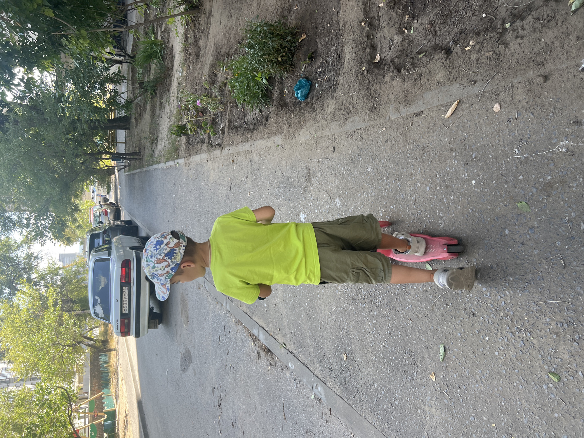 A young boy wearing a lime green t-shirt, olive green cargo shorts, a patterned baseball cap, and grey sneakers is riding a pink scooter on a paved sidewalk. The boy is viewed from behind as he travels along the sidewalk, which runs parallel to a street with several parked cars, including a white sedan with the license plate "C533YE34". The sidewalk is next to a dirt area with some sparse vegetation and a blue plastic bag on the ground. Trees line the far side of the street, and a building is visible in the background. The scene appears to be a residential street in Volgograd, Russia.
