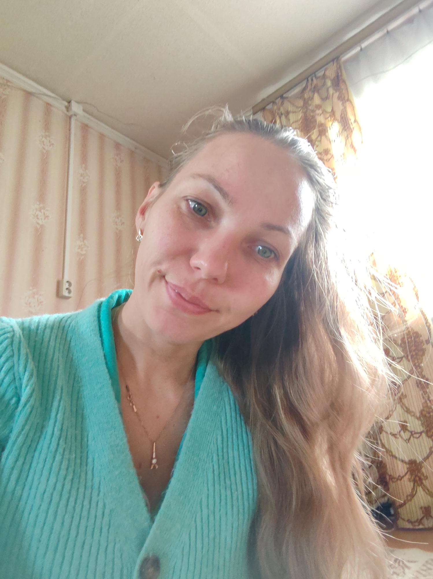 This is a close-up, slightly angled selfie of a woman indoors, likely in Чухлэм, Russia. She has long, light brown hair, which falls over her shoulders, and striking green eyes. Her head is tilted slightly, and she offers a gentle, natural smile. She wears a ribbed, teal-colored cardigan over a similar top, and a delicate gold necklace with a small pendant shaped like the Eiffel Tower. A small, square, silver-colored earring is visible in her right ear.
The background suggests a simple, domestic setting. The left wall is covered with light-toned wallpaper featuring a subtle, repeating pattern. A white electrical conduit runs along the top edge of this wall, and a Type F electrical outlet is mounted on it. The ceiling is white. To the right, intense natural light streams in from a window, which is partly obscured by a curtain with a warm-toned, intricate pattern in brown and cream. The bright illumination indicates it is daytime. The woman appears calm and content. No visible text is present in the image.