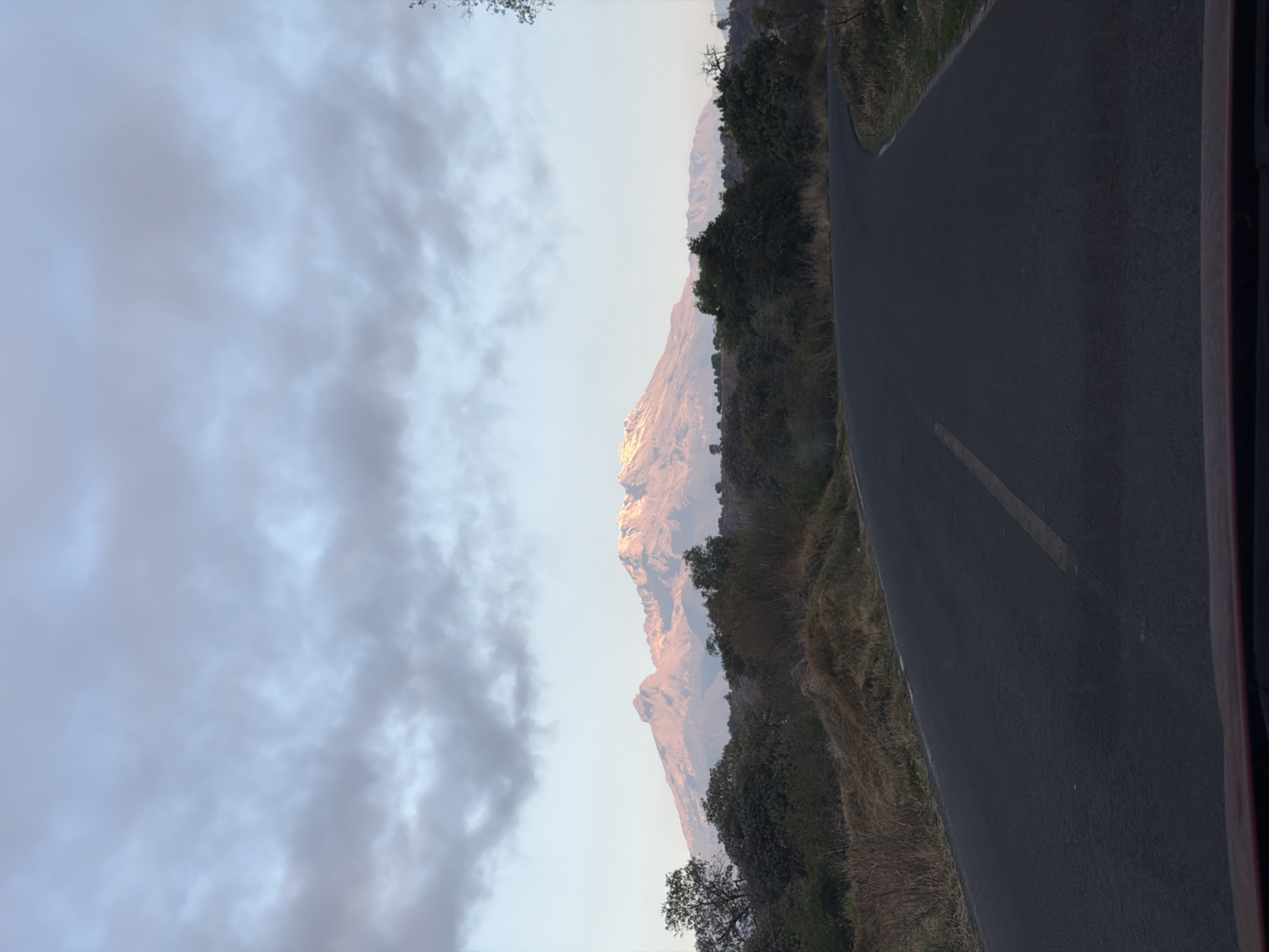 The image shows a road next to a mountain in Santiago Xalitzintla, Mexico. The road is asphalt and has a yellow line. The mountain is rocky and covered with vegetation. The sky is cloudy.