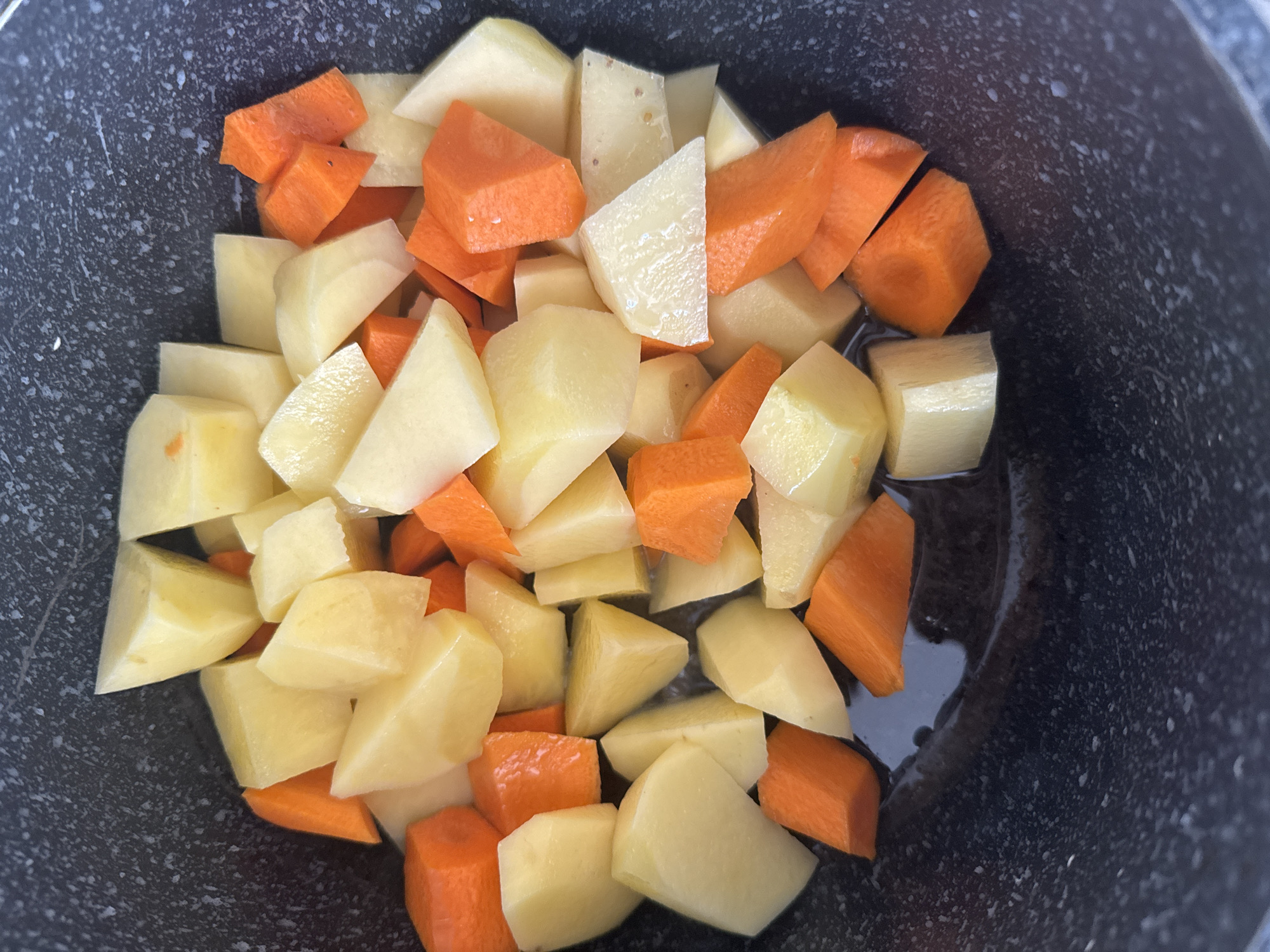 A top-down, close-up shot shows a dark pot filled with chopped potatoes and carrots in some liquid. The potatoes are cut into roughly uniform, irregular chunks, displaying a pale yellow flesh and white peel. The carrots are also cut into similar-sized pieces, showcasing their bright orange color. The vegetables are mixed together, creating a colorful pattern against the dark background of the pot, which has a speckled texture. The liquid at the bottom of the pot is dark and appears to be water or broth. The scene is devoid of human presence and any recognizable city landmarks.
