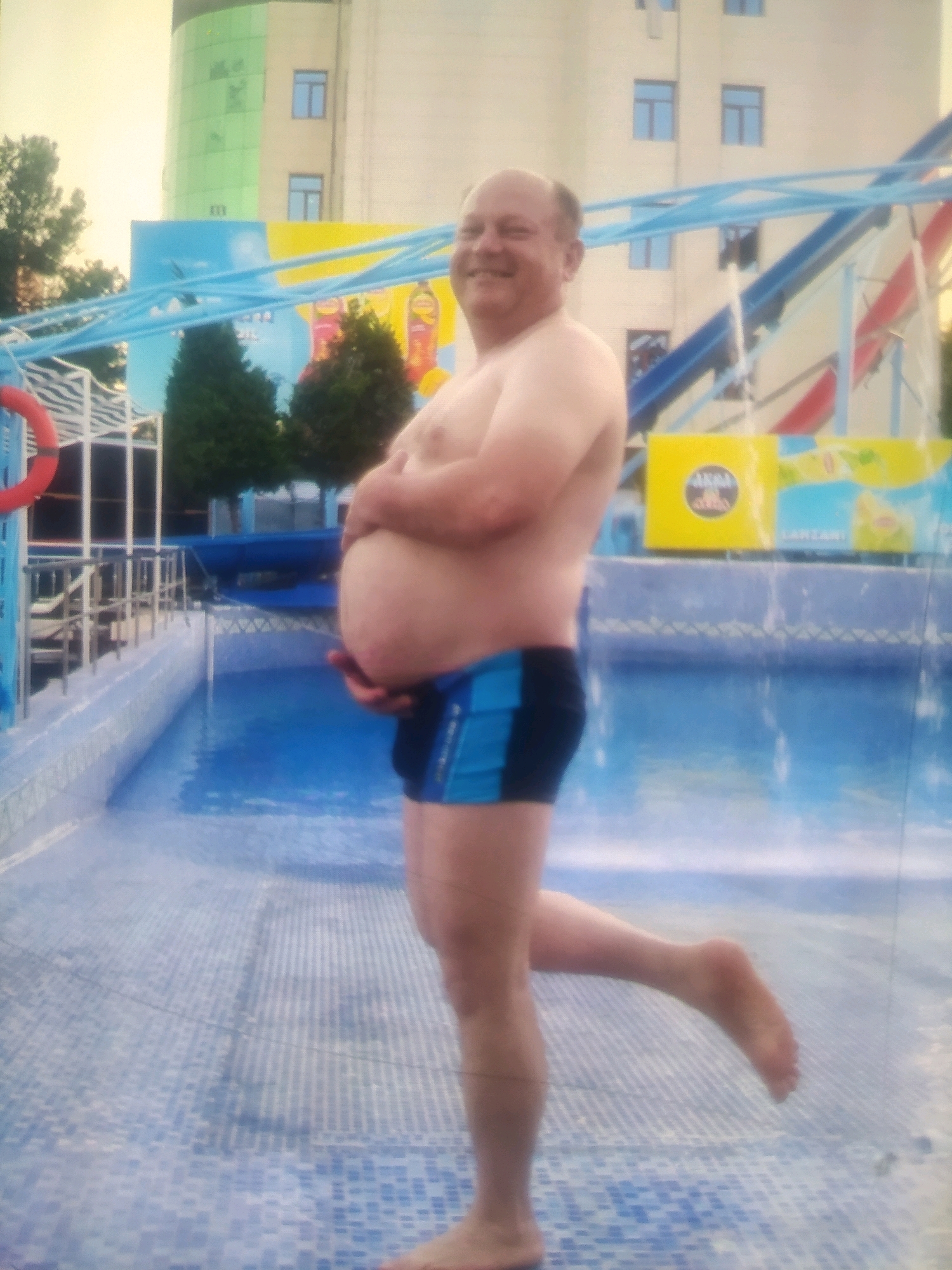 A man stands by a swimming pool in Birlik, Uzbekistan. He is facing to the left and is wearing blue and black swimming trunks. He has a visible stomach. He is smiling at the camera and has his right leg slightly raised. In the background, there is a water slide and a building with green and beige panels.