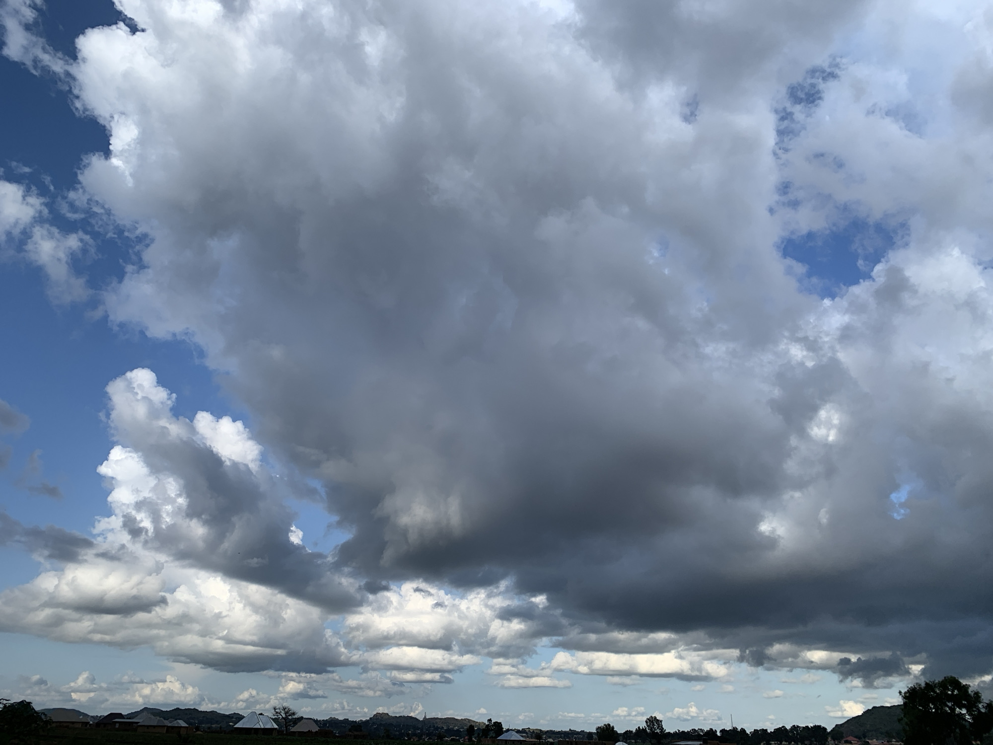 The image captures a broad, overcast sky over Kuru Karama, Nigeria. The sky is dominated by large, grey, and white clouds, with patches of blue sky visible. The clouds appear dense and possibly indicative of impending rain. In the lower portion of the image, a flat landscape is visible with scattered buildings and trees. The architecture of the buildings suggests a rural or semi-urban setting, with some structures featuring conical roofs. The overall scene conveys a natural environment under a dramatic sky.