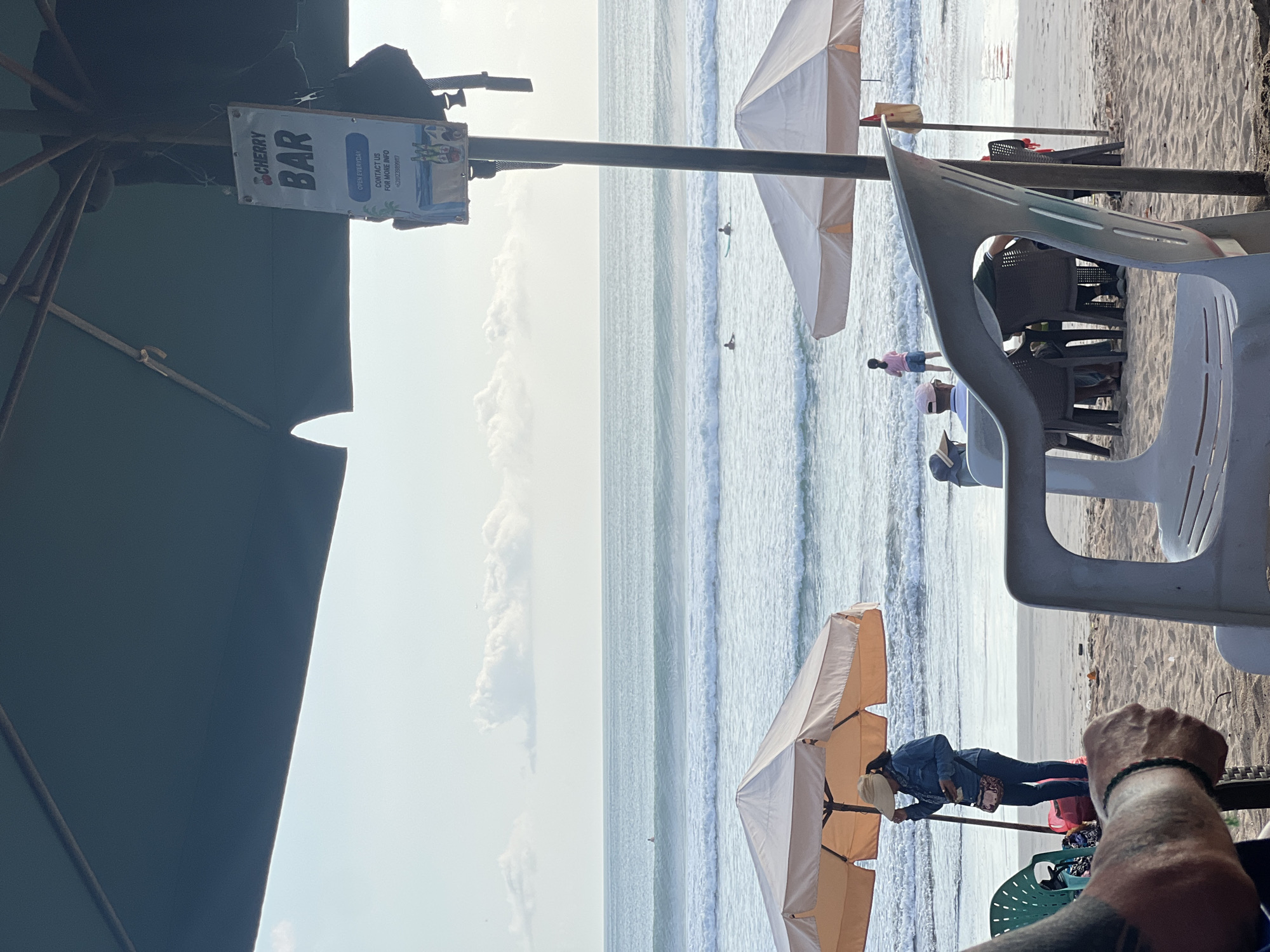 This image depicts a scene on Kuta Beach in Kuta, Indonesia, on a hazy day with calm seas and gentle waves. In the foreground, a white plastic chair faces away from the camera, and to the left, a tattooed arm with a green bracelet is visible. Further back, a woman in a denim jacket and hat stands under a peach-colored umbrella, looking at her phone. Several tables and chairs are set up on the sand, some occupied by people facing the ocean. Two white umbrellas provide shade on the beach. In the background, several surfers are visible in the water, and a sign for "CHERRY BAR" hangs from a pole, indicating the presence of a beach bar. The sky is overcast with scattered clouds.