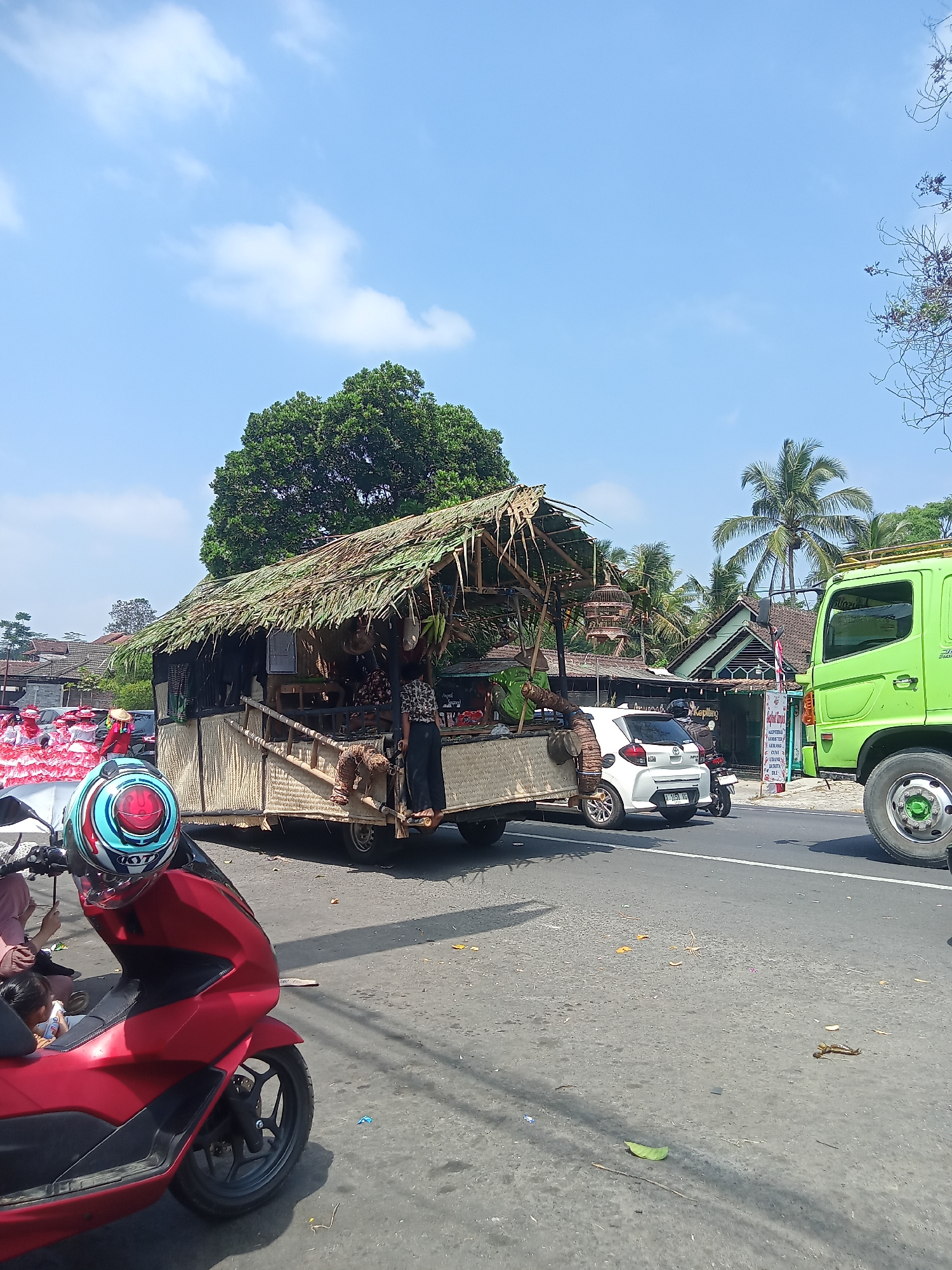 The image depicts a street scene in Urutsewu, Indonesia. In the foreground, a red motorcycle is parked, its front wheel and handlebars visible. A helmet with blue, red, and white accents sits on the handlebars. Behind the motorcycle, a flatbed trailer is being pulled, constructed with woven material and topped with a thatched roof, resembling a small hut or stall. Two individuals are visible on or near the trailer. To the right of the trailer is a white car. Further to the right, a large green truck is stopped on the road. In the background, a line of people in red and white attire are visible, suggesting a parade or procession. Palm trees and other vegetation are present, with buildings and a large tree in the middle ground. The sky is blue with scattered white clouds. The scene appears to be a sunny day.