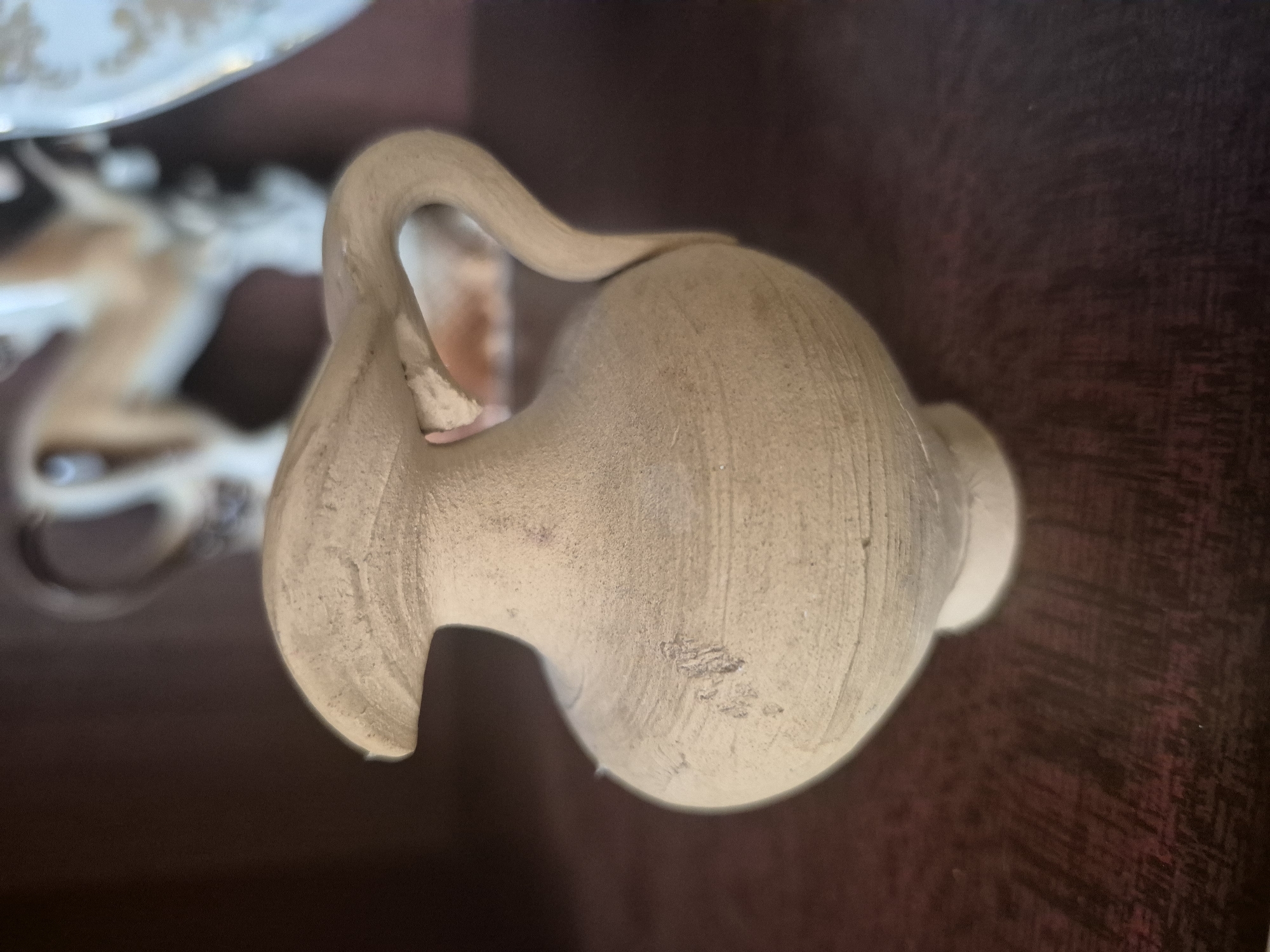 A close-up, eye-level shot shows a small, light-brown, unglazed ceramic jug with a wide lip and a curved handle. The jug sits on a dark, wooden surface, likely a shelf. In the blurred background, other objects can be seen, including what appears to be a decorative porcelain plate with gold accents and a light-colored statue of an animal, possibly a deer or goat. The lighting is dim, suggesting an indoor setting. The image was taken in Vanadzor, Armenia.