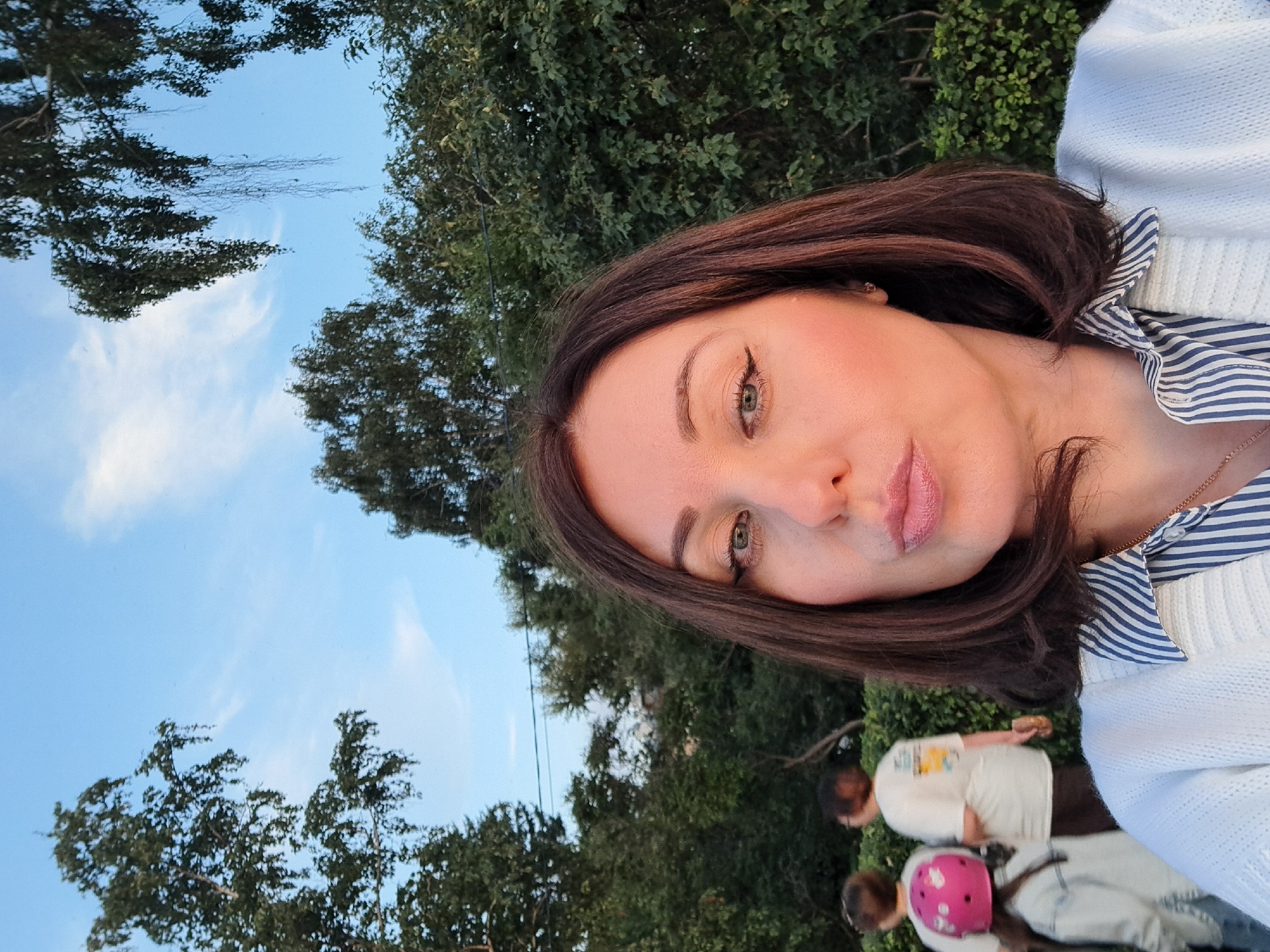 A woman is taking a selfie outdoors, with a background of trees and a blue sky. She has medium-length brown hair and is wearing a white cardigan over a striped shirt. Her lips are slightly pursed. In the blurred background, two people are visible, one wearing a white t-shirt and the other a white t-shirt and jeans with a pink helmet. The location is the city of Irkutsk, Russia.