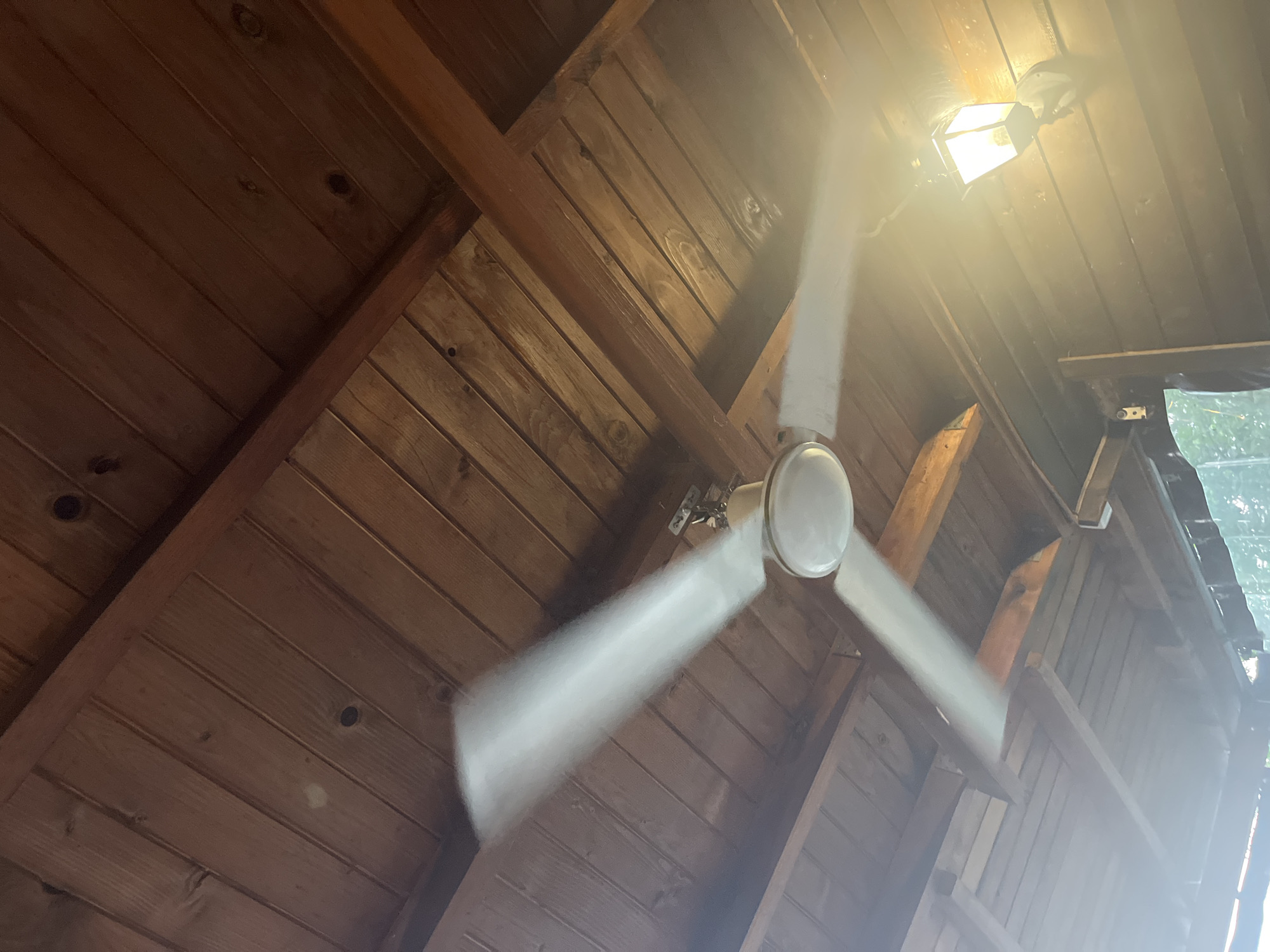 The scene depicts an upward-angled view within a sheltered wooden structure in Varshilo, Bulgaria. The ceiling is constructed from reddish-brown wooden planks arranged linearly, with prominent dark knots and grain patterns, and reinforced by larger, darker wooden beams. A three-bladed white ceiling fan, suspended from the wooden ceiling, is actively rotating; its blades are rendered as blurred streaks due to motion. The fan's central housing includes a circular light fixture. To the lower right, a wall-mounted lantern with a dark frame and glass panels illuminates the area, casting a bright, warm light and generating significant lens flare. The wall behind the lantern is also planked wood. The bottom left edge of the image provides a blurred glimpse of green foliage, indicating an outdoor or semi-outdoor setting beyond the wooden structure. No human or animal subjects are present.