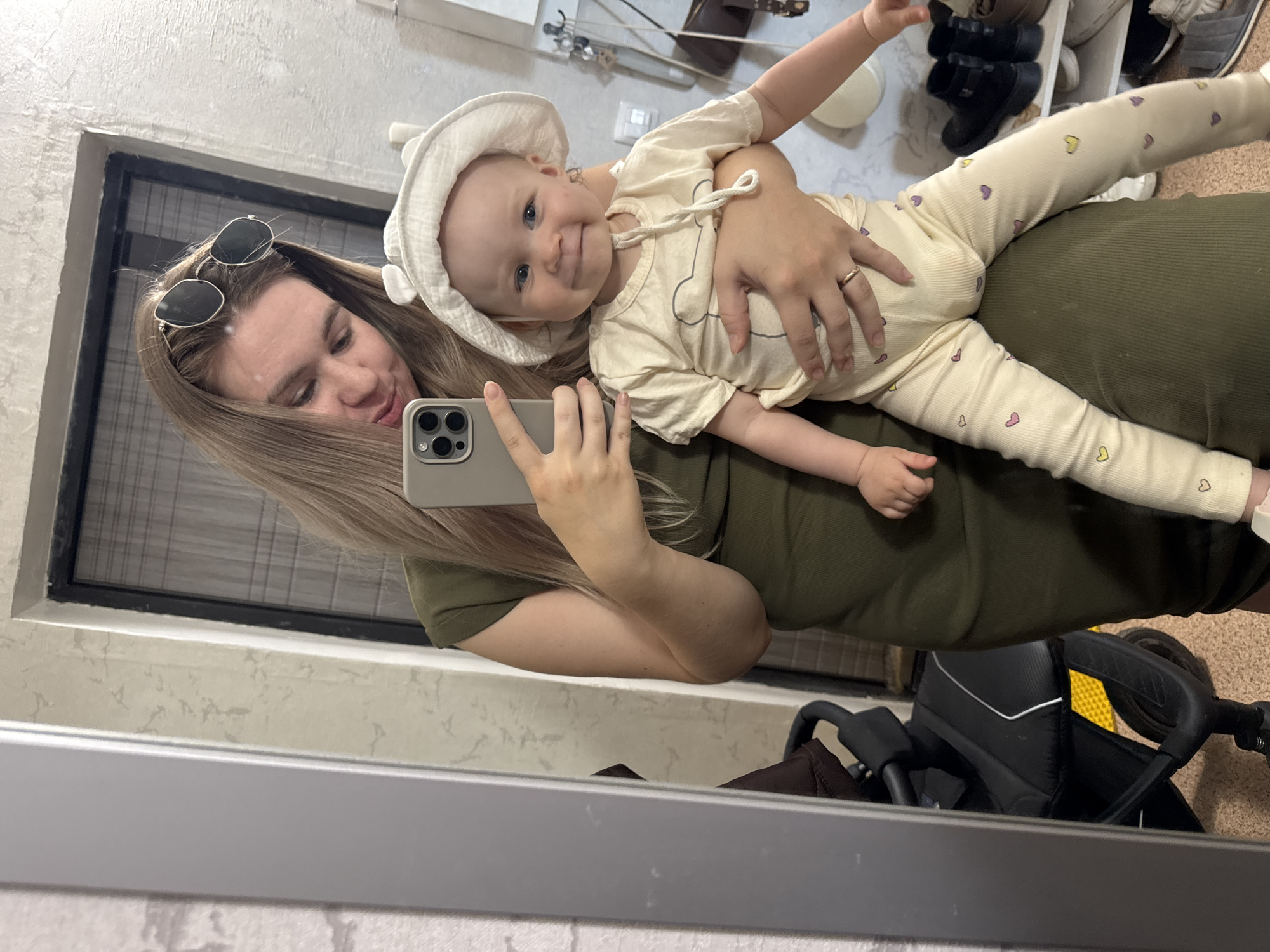 A mirrored reflection captures a woman holding a baby. The woman has light brown hair, wears sunglasses on her head, and is holding a phone to take a picture of herself and the baby. She is wearing an olive-green outfit. The baby, wearing a light-colored outfit with heart details, also has a white hat. The reflection also shows a baby stroller and a shelf with shoes. The location appears to be indoors, possibly a residence, in Chaikovsky, Russia.