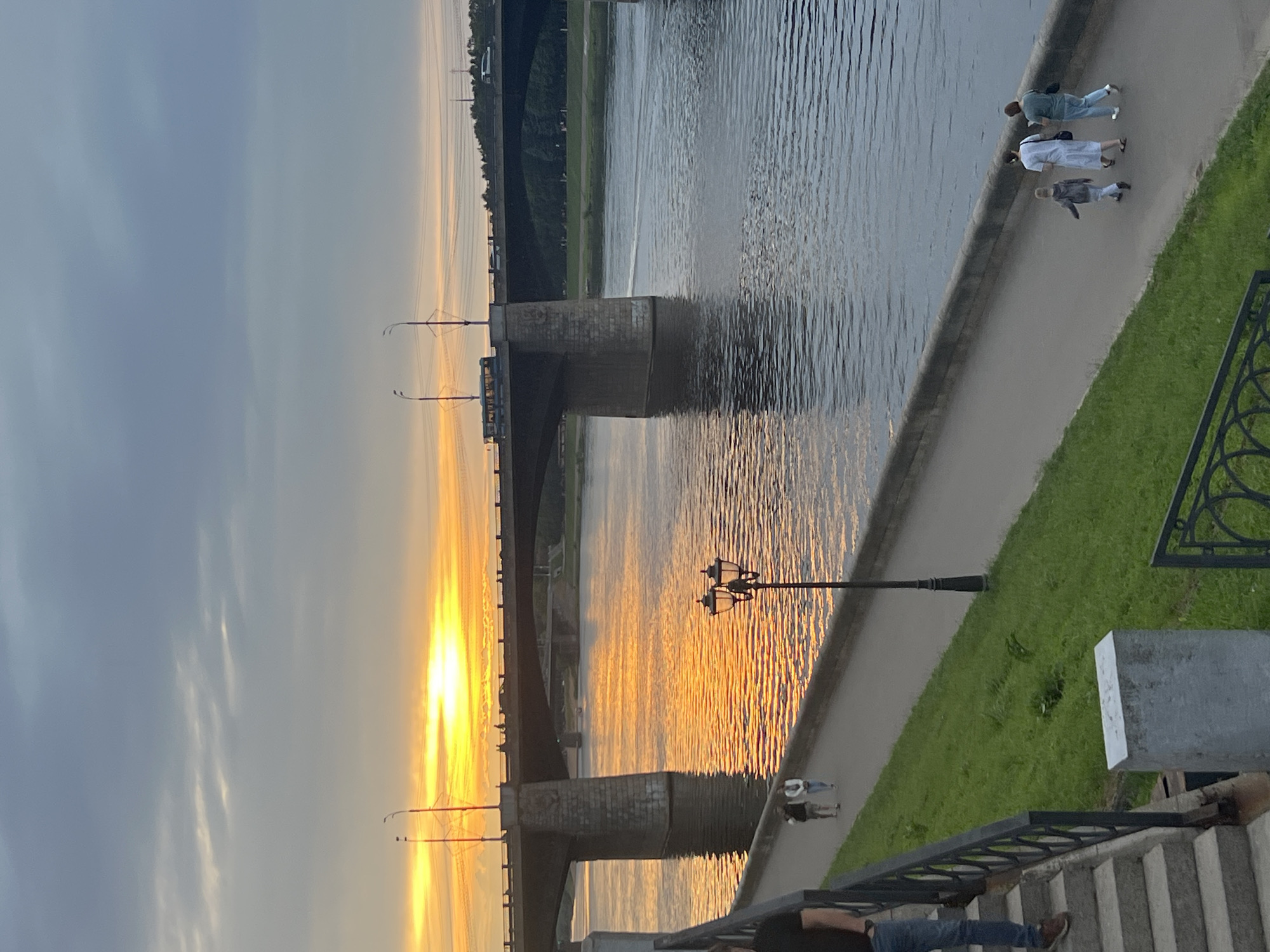 This image depicts a view of the Volga River in Tver, Russia, at sunset. A large stone bridge spans the river, with its reflection shimmering in the water. The sky is filled with dramatic clouds, illuminated by the setting sun. On the riverbank, a paved walkway runs alongside the water, bordered by a strip of green grass and a stone wall. Several people are visible walking along the path, including a family with a child. A street lamp stands on the walkway, casting a warm glow. In the foreground, a set of stone stairs leads down to the walkway, with a person standing on them. The overall scene is serene and picturesque, capturing the beauty of a summer evening in Tver.