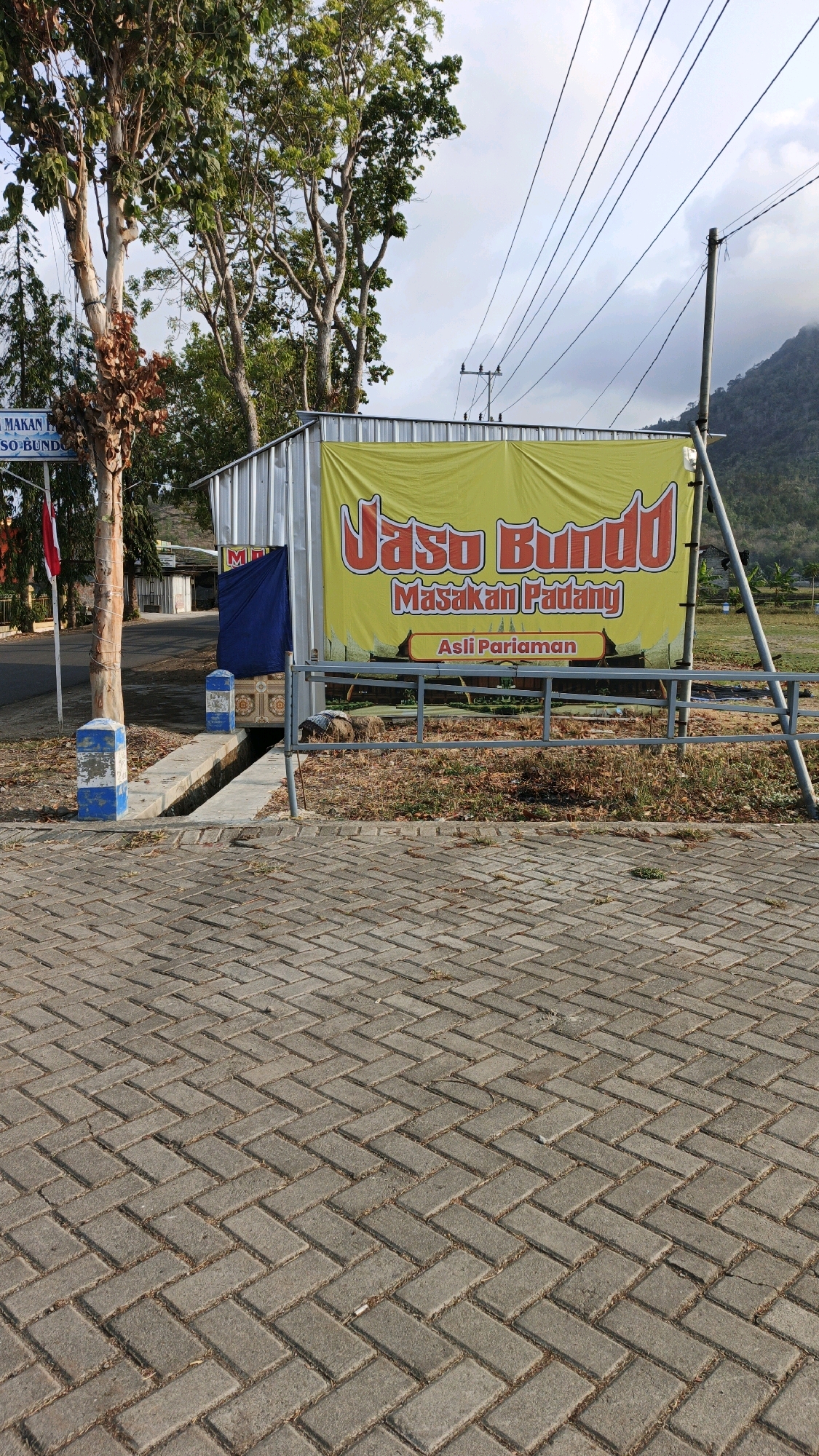 In Tanggulwelahan, Indonesia, a street-level view captures a yellow banner prominently displayed on a metal-roofed structure. The banner reads "Jaso Bundo Masakan Padang" with the subtitle "Asli Pariaman". To the left of the structure, a red and white flag flies on a pole. The foreground is paved with gray interlocking bricks laid in a herringbone pattern. Behind the banner, dry vegetation and a grey metal railing are visible. The background shows trees, utility poles with overhead wires, and a hilly landscape under a cloudy sky.