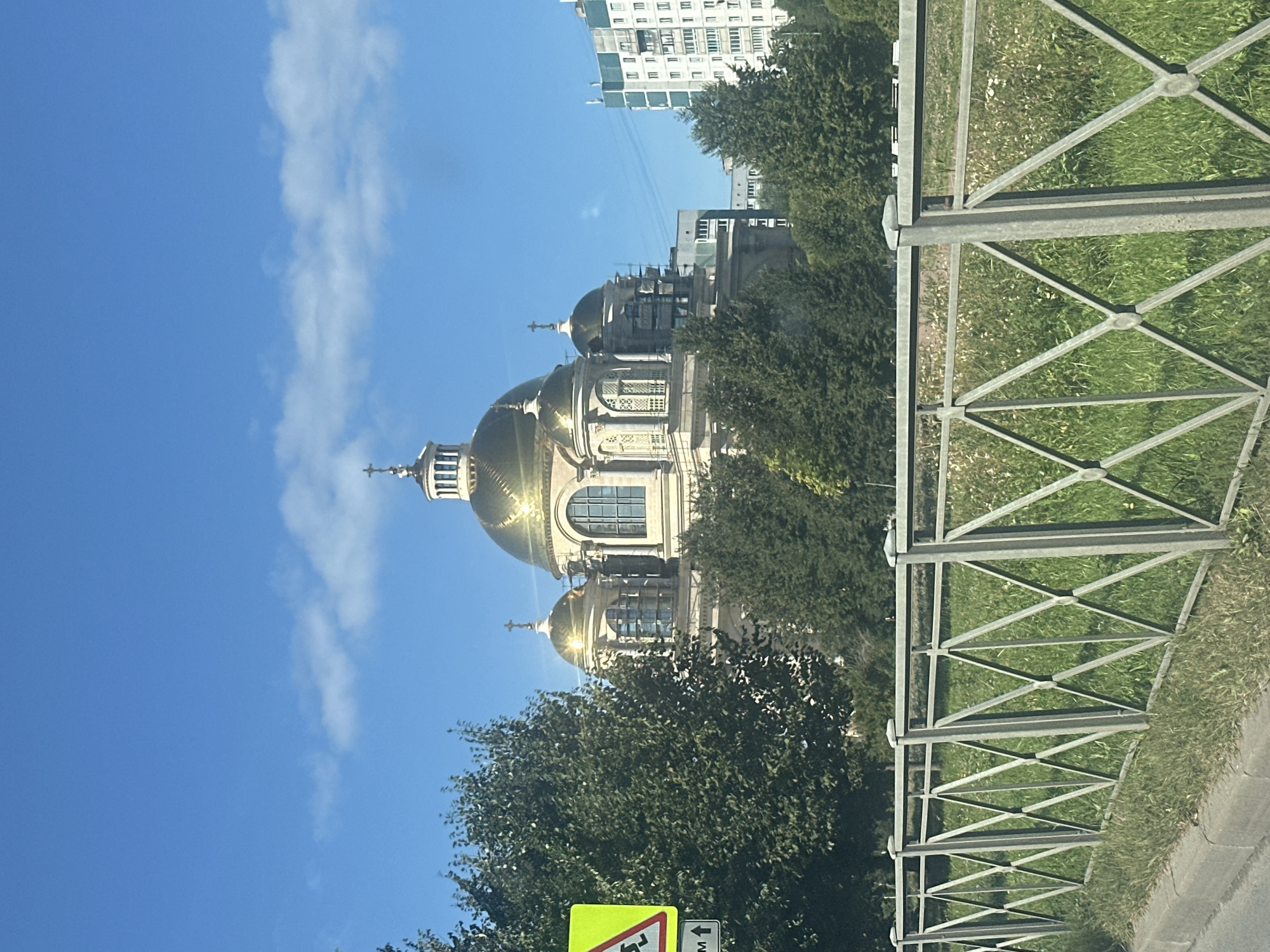 The image captures a sunny day scene with a grand golden-domed church as the central subject. The church, possibly the Saint Isaac's Cathedral or Kazan Cathedral given the architectural style and golden domes, is located in Saint Petersburg, Russia. Its imposing structure features multiple smaller domes and a large central dome topped with a cross. The church is surrounded by lush green trees and some scaffolding can be observed on its side, suggesting ongoing maintenance or construction. In the foreground, a grey metal fence with a diamond pattern runs horizontally across the image, separating a sidewalk from a grassy area with scattered small white flowers. A yellow triangular road sign depicting a running child is visible on the left, indicating a pedestrian crossing or school zone. Behind the church, a modern residential building with multiple windows and balconies is visible against a clear blue sky with a few wispy clouds. The overall impression is one of a serene urban landscape with historical and modern elements coexisting.