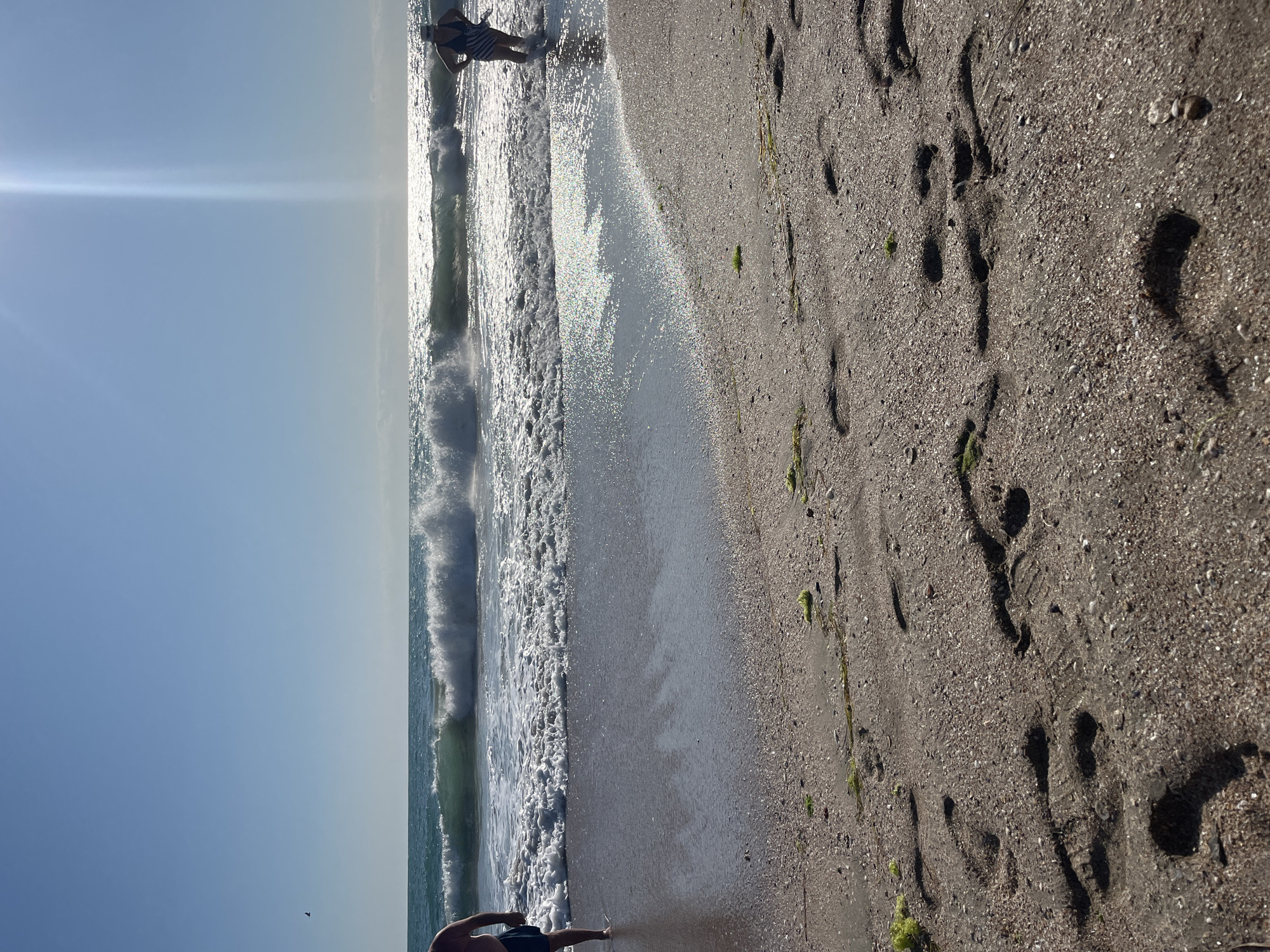 The image captures a sunny day at Pomorie Beach in Bulgaria. The foreground is dominated by wet sand with numerous footprints, suggesting recent activity. Some green seaweed is scattered on the sand. The middle ground shows gentle waves breaking on the shore, with the water sparkling under the bright sun. Two people are visible. On the left, a person stands in the shallow water, facing away from the camera. Their lower body is visible, wearing dark blue shorts. On the right, another person is standing in the water, also facing away, wearing a striped swimsuit and a cap. The background consists of the sea under a clear blue sky. A prominent sunbeam pierces the sky and reflects off the water, creating a bright glare. In the far distance, a single bird is flying in the sky.