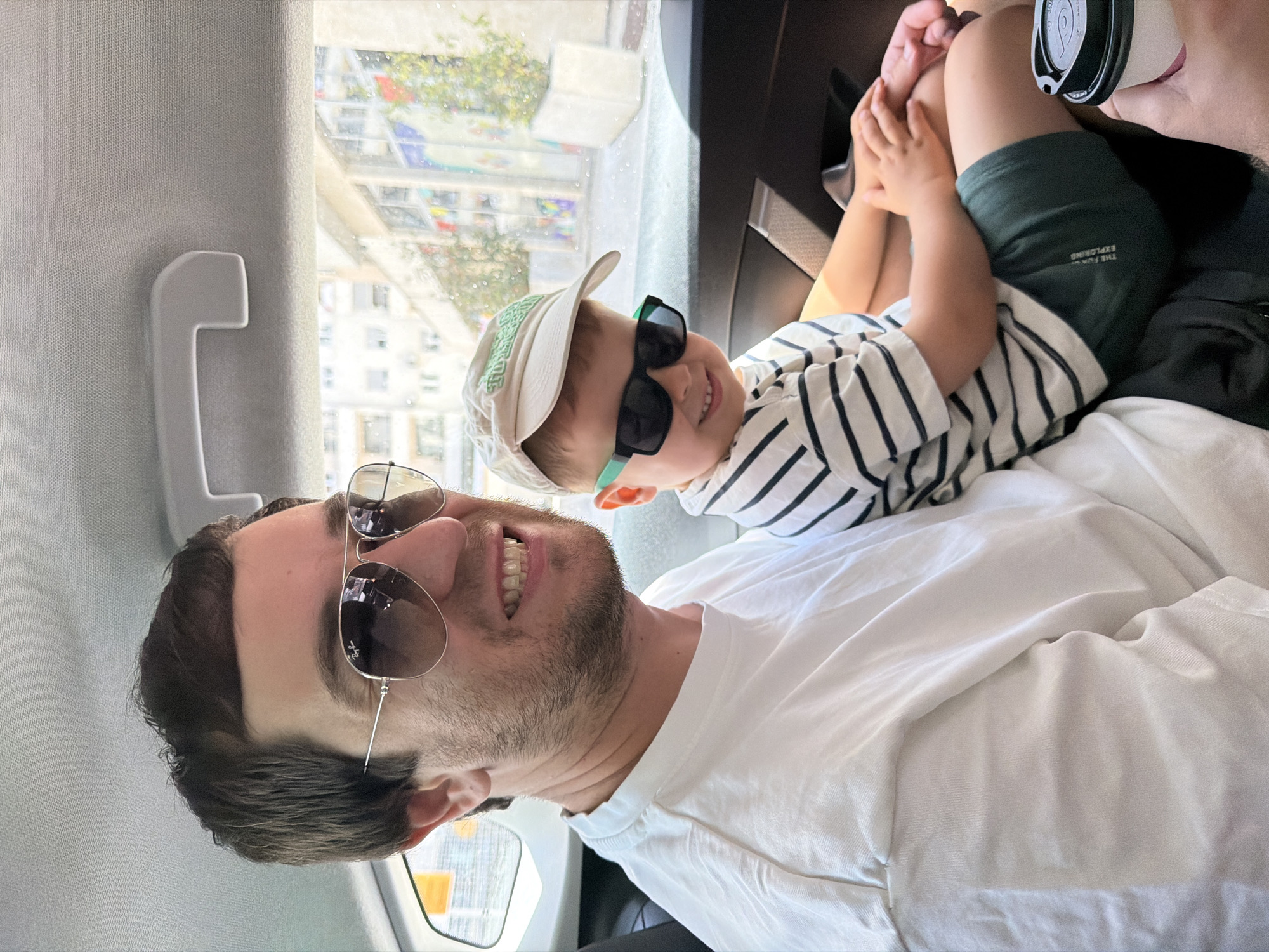A man and a young child are captured in a bright, selfie-style photograph taken inside a vehicle, likely a car or bus, in Budva, Montenegro. The image is oriented sideways, with both subjects looking towards the camera.
The man, in the foreground, has dark hair and stubble. He wears a white crew-neck t-shirt and aviator sunglasses, with a visible "Ray-Ban" logo on the lens. He is smiling broadly, showing his teeth.
A young child is positioned slightly behind and to the side of the man, possibly on his lap. The child wears a light-colored baseball cap and dark, child-sized sunglasses with green frames. The child's attire includes a white t-shirt with dark horizontal stripes and dark green shorts. Visible text on the child's shorts reads "THE FUN EXPLORING." The child is also smiling.
Through the vehicle's window in the background, a cityscape with buildings and some greenery is visible, indicating an urban or suburban environment. The bright natural light suggests it is daytime and sunny. A white grab handle is present on the interior side panel of the vehicle. Part of a cup with a lid is also visible near the child's hands. Both subjects appear happy and content.