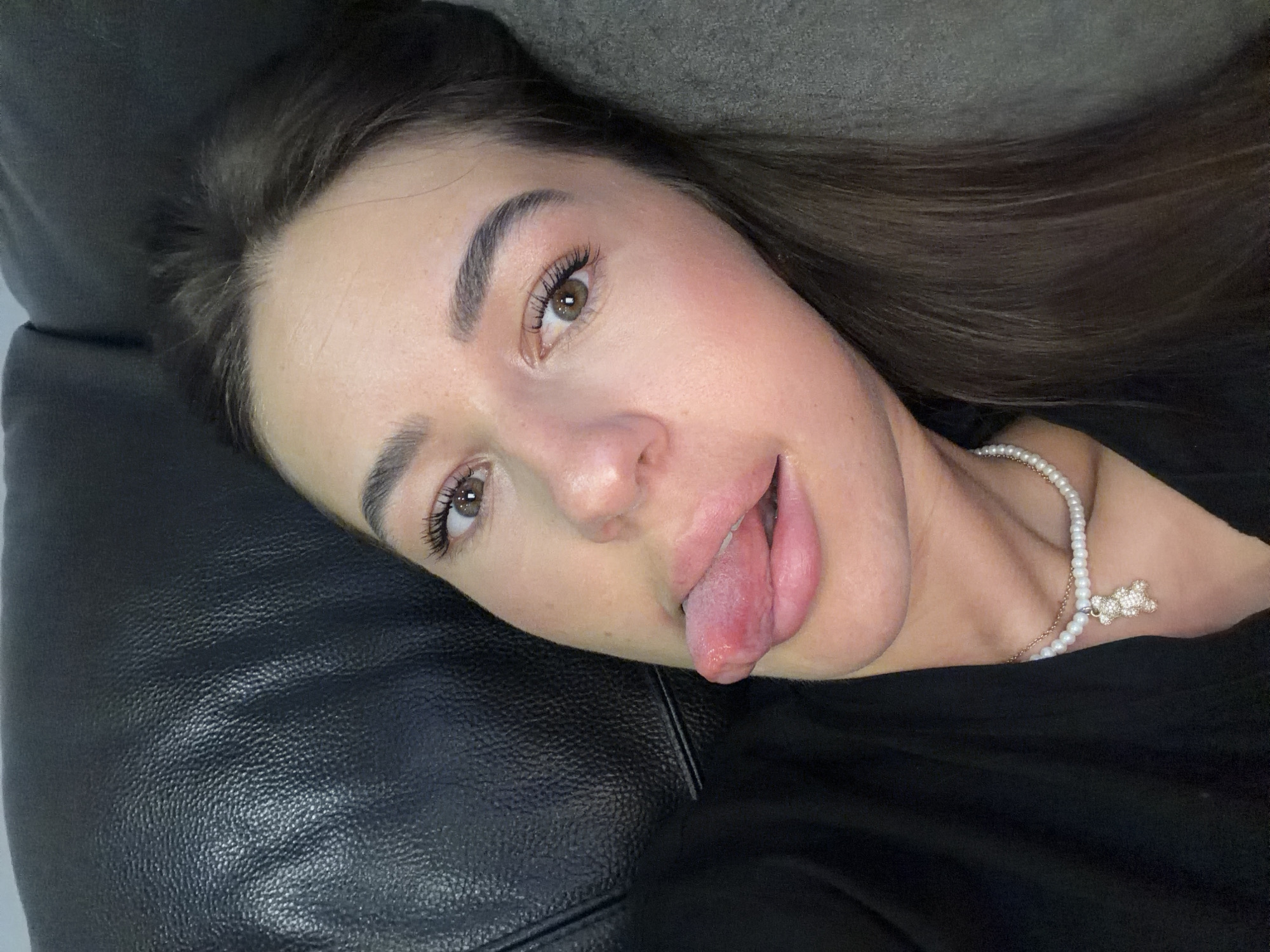 The image is a close-up, selfie-style photograph of a young woman lying down, possibly on a couch, in what appears to be a domestic setting in Novi Iskar, Bulgaria. She is wearing a black top and a pearl necklace with a small, sparkling teddy bear pendant. Her hair is dark brown and falls around her face and shoulders. The woman is sticking her tongue out, and her mouth is slightly open. Her expression is playful or mischievous. The background is dominated by a dark, textured surface, likely a leather couch, and a darker, softer surface, possibly a pillow or blanket. The lighting is soft, highlighting her face and features.