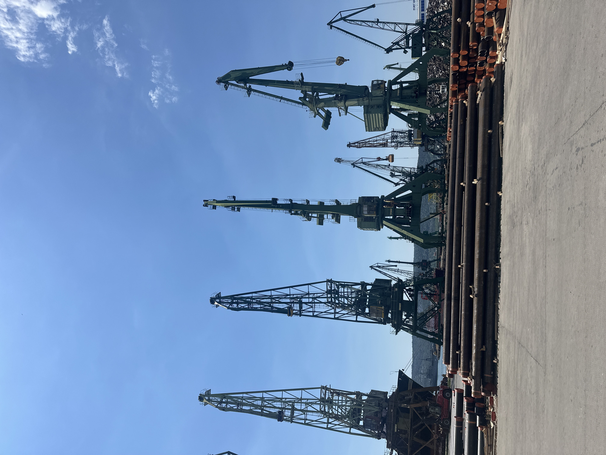 This image shows a harbor scene with multiple cranes of varying sizes and designs silhouetted against a clear blue sky with some wispy white clouds. In the foreground, a paved area is visible to the right, and a stack of large metal pipes runs along the left side of this paved area. Behind the pipes and cranes, there appears to be a body of water and a hilly landscape in the distance.
The cranes are the dominant objects in the scene, suggesting an industrial or port environment where heavy lifting and loading operations take place. Some of the cranes are massive, industrial structures made of metal lattice, while others appear to be smaller, more modern-looking cranes. The overall impression is one of a busy working port.
The lighting suggests it is daytime, likely morning or afternoon, given the bright sunlight. The clear sky indicates fair weather. There is no visible text in the image. The scene evokes a sense of industry and productivity, with the imposing cranes standing as symbols of commerce and trade.