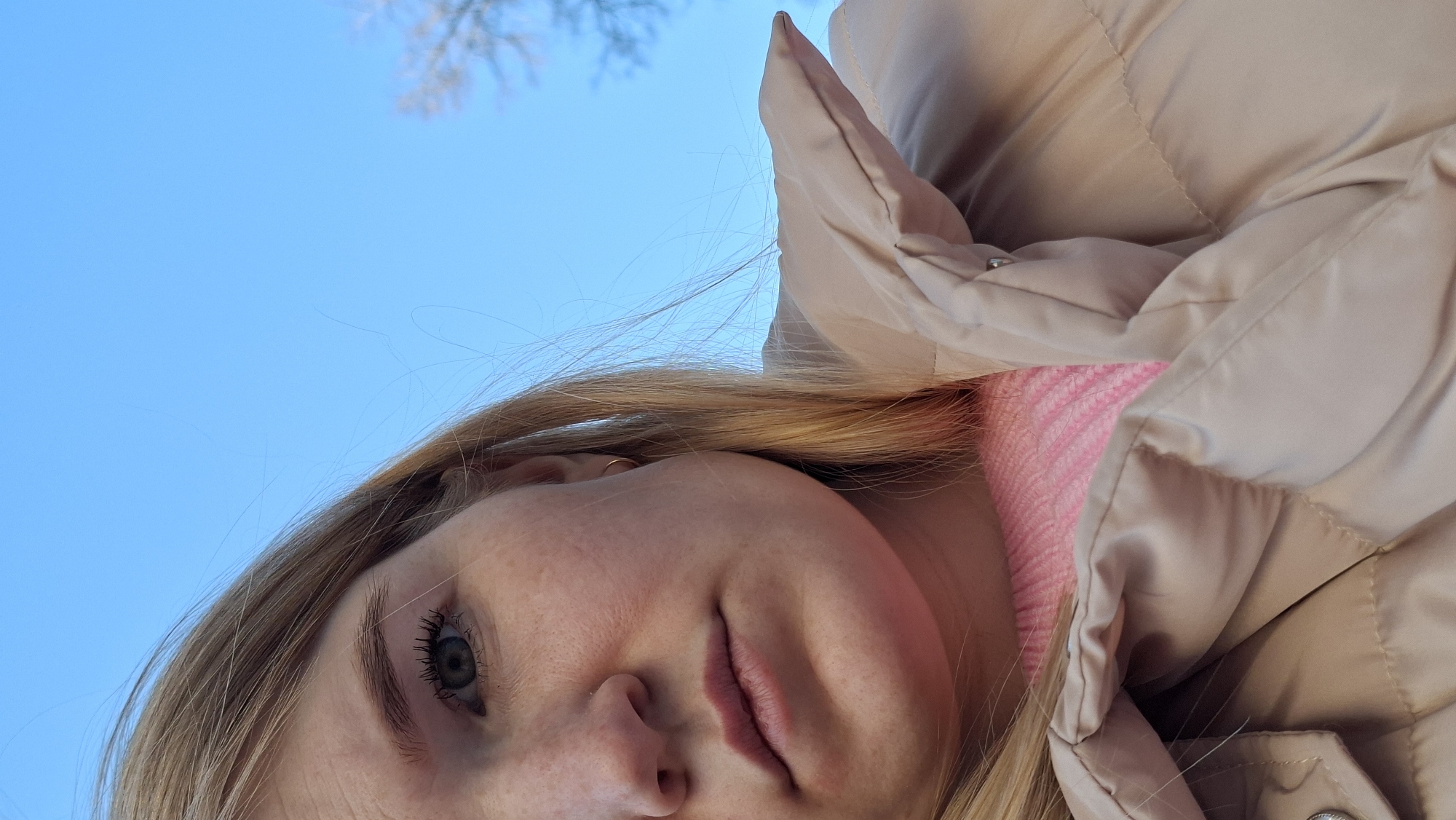 A close-up, low-angle portrait captures a young woman looking down towards the camera, set against a vibrant blue sky. Her face, positioned in the lower left quadrant, reveals light blue or grey eyes with visible mascara on her eyelashes, subtle freckles across her nose and cheeks, and naturally colored lips, conveying a calm expression. Her long, blonde hair frames her face, with some strands appearing wispy against the sky.
She is dressed in winter clothing, featuring a light beige or tan puffer jacket that partially covers her head and shoulders, and a bright pink knitted sweater visible underneath. These garments suggest cold weather conditions.
The background is dominated by a clear, bright blue sky, indicating daytime. In the upper right corner, the blurred, bare branches of a tree are faintly discernible, consistent with late autumn, winter, or early spring in Chaikovsky, Russia. The camera's upward tilt creates a dynamic perspective, as if she is being photographed from below while gazing upwards. No text is visible in the image.