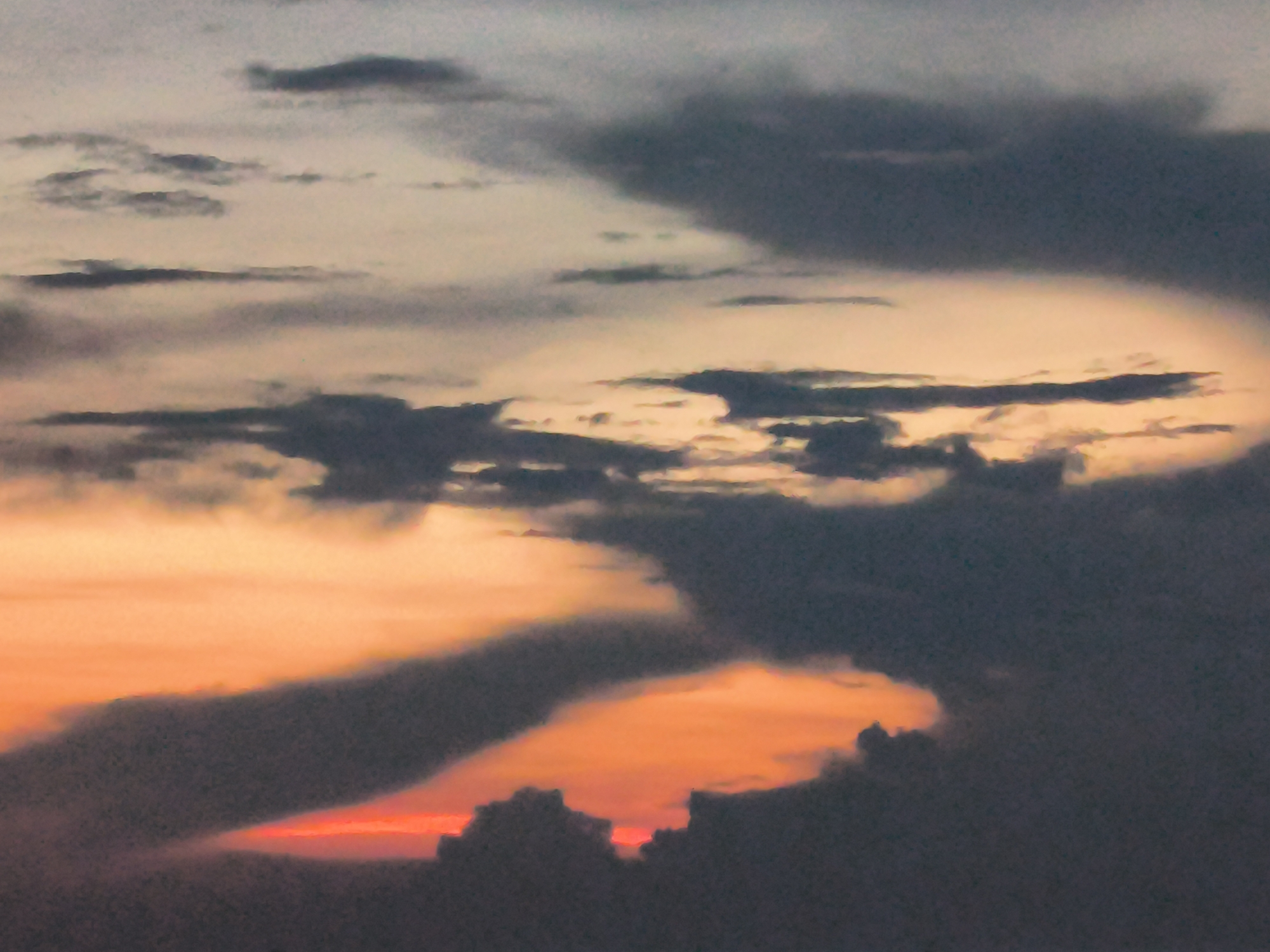 The image captures a dramatic sky at dusk in Lhokseumawe, Indonesia. The sky is filled with dark, heavy clouds, some tinged with orange and red light from the setting sun, which is partially obscured by the cloud cover. The overall environment suggests a potentially stormy or overcast evening.