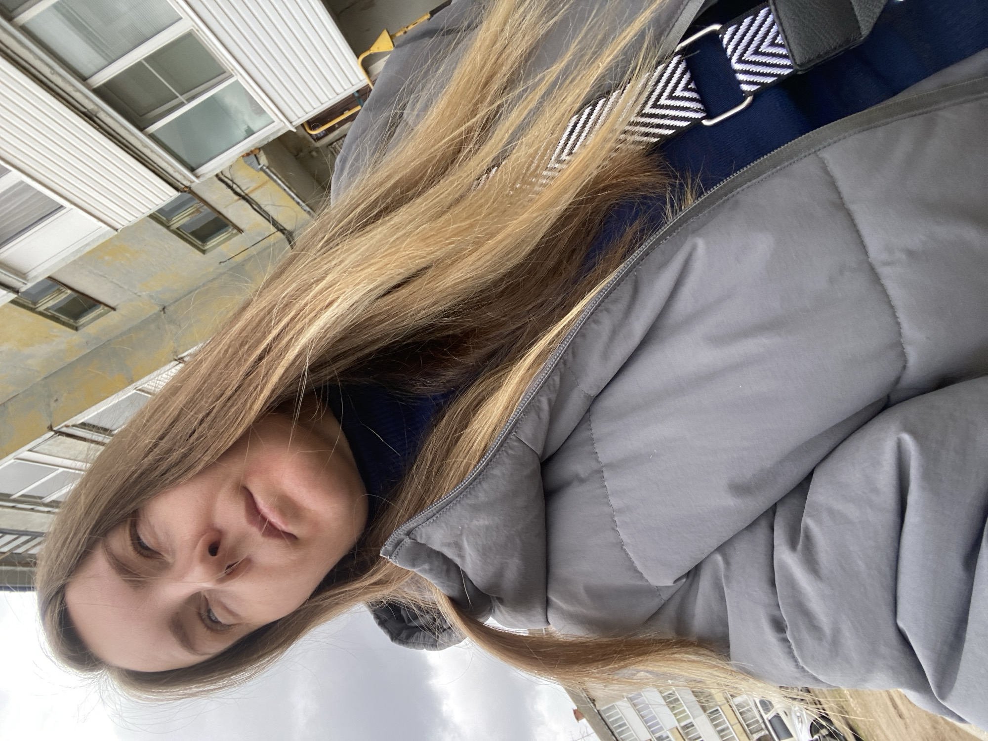 The image is a vertical portrait of a young woman, likely a selfie, captured against an urban backdrop in Noviy, Russia. She is positioned slightly to the left of the frame, looking forward with a neutral, calm expression. Her long, straight, light brown hair cascades over her shoulders and body.
She is dressed in a grey puffer jacket, partially zipped, beneath which a dark blue garment is visible. A prominent detail is the black and white chevron-patterned strap of a bag, secured by a silver buckle, draped over her right shoulder.
The background features the facade of a building, characterized by light-colored, weathered walls with dark stains and multiple rectangular windows. Portions of white, ribbed siding are also visible on the structure. The upper part of the image reveals an overcast, grey sky, suggesting a cloudy daytime. No text is visible in the scene.