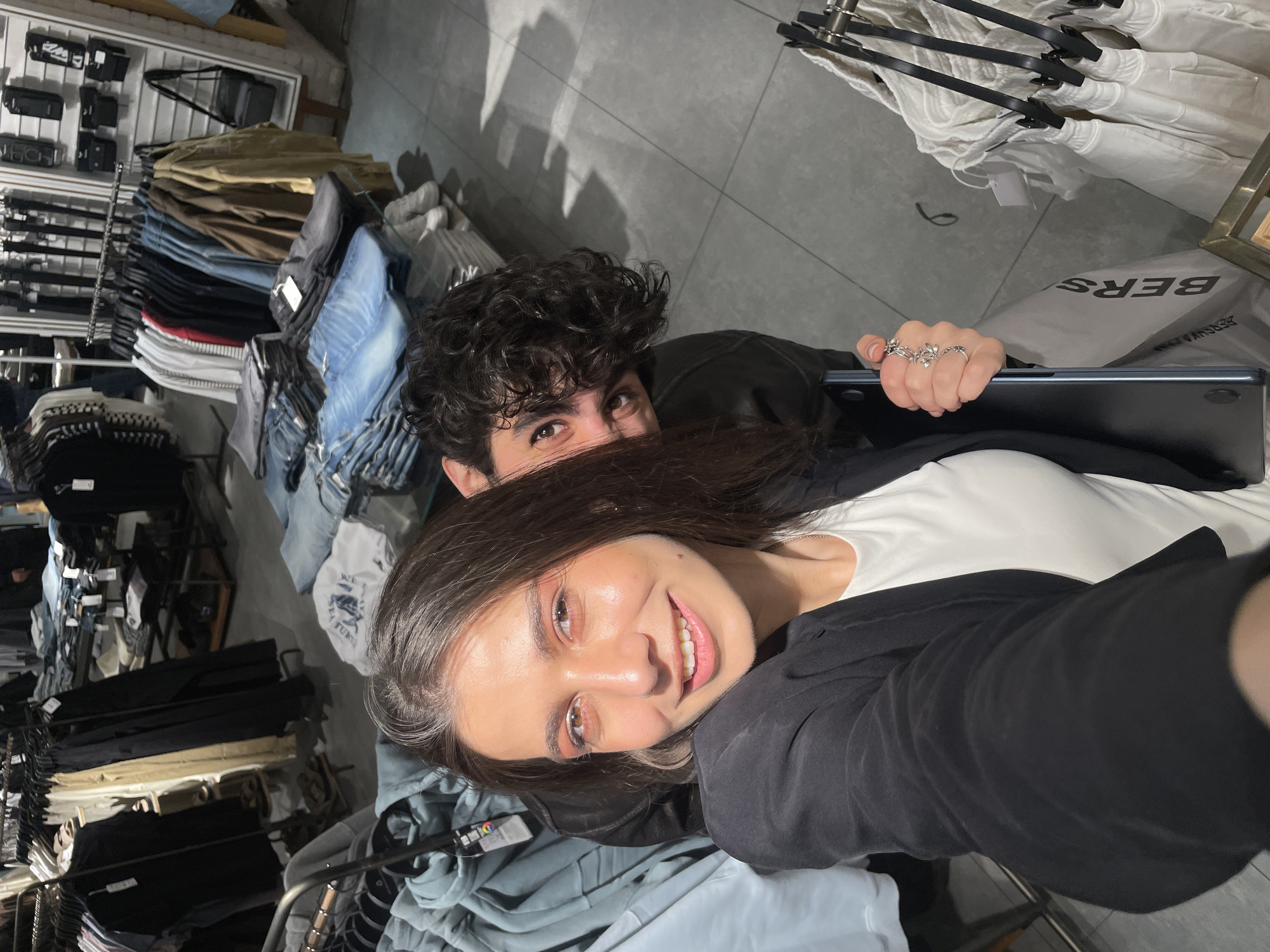 Two people, a woman and a man, are taking a selfie. The woman is in the foreground, smiling with her face turned towards the camera. She has long brown hair and is wearing a white top under a black jacket. The man is positioned behind her, partially obscured by her hair, but visible with his curly dark hair and brown eyes.
They appear to be inside a clothing store. Clothing racks are visible on both sides of the frame, displaying various items such as jeans, shirts, and jackets. In the upper right corner, there is a visible "BERSHKA" logo on a shopping bag. The woman's hand is holding a black laptop. The lighting suggests an indoor retail environment. The location is Yerevan, Armenia, inferred from the presence of a Bershka store, which is a global retail chain with a presence in Yerevan.