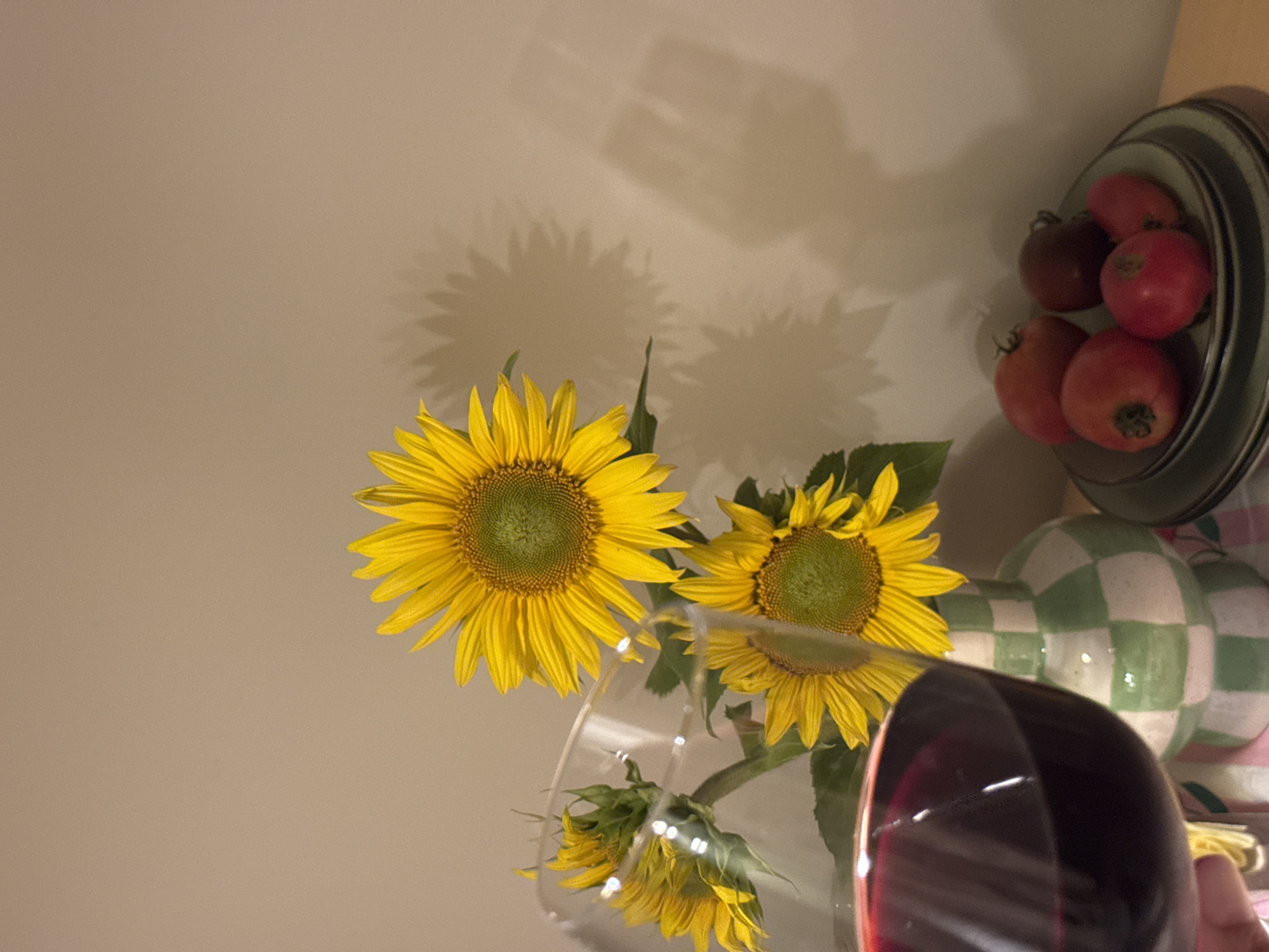 A close-up shot captures a glass of red wine in the foreground, with sunflowers and a stack of plates containing tomatoes in the background. The sunflowers are yellow with green centers, and they are arranged in a checkered vase. The tomatoes are red and various sizes. The scene appears to be set indoors with a plain wall as the backdrop. The lighting creates shadows of the sunflowers on the wall. There is no identifiable information to suggest a location in Izhevsk, Russia.