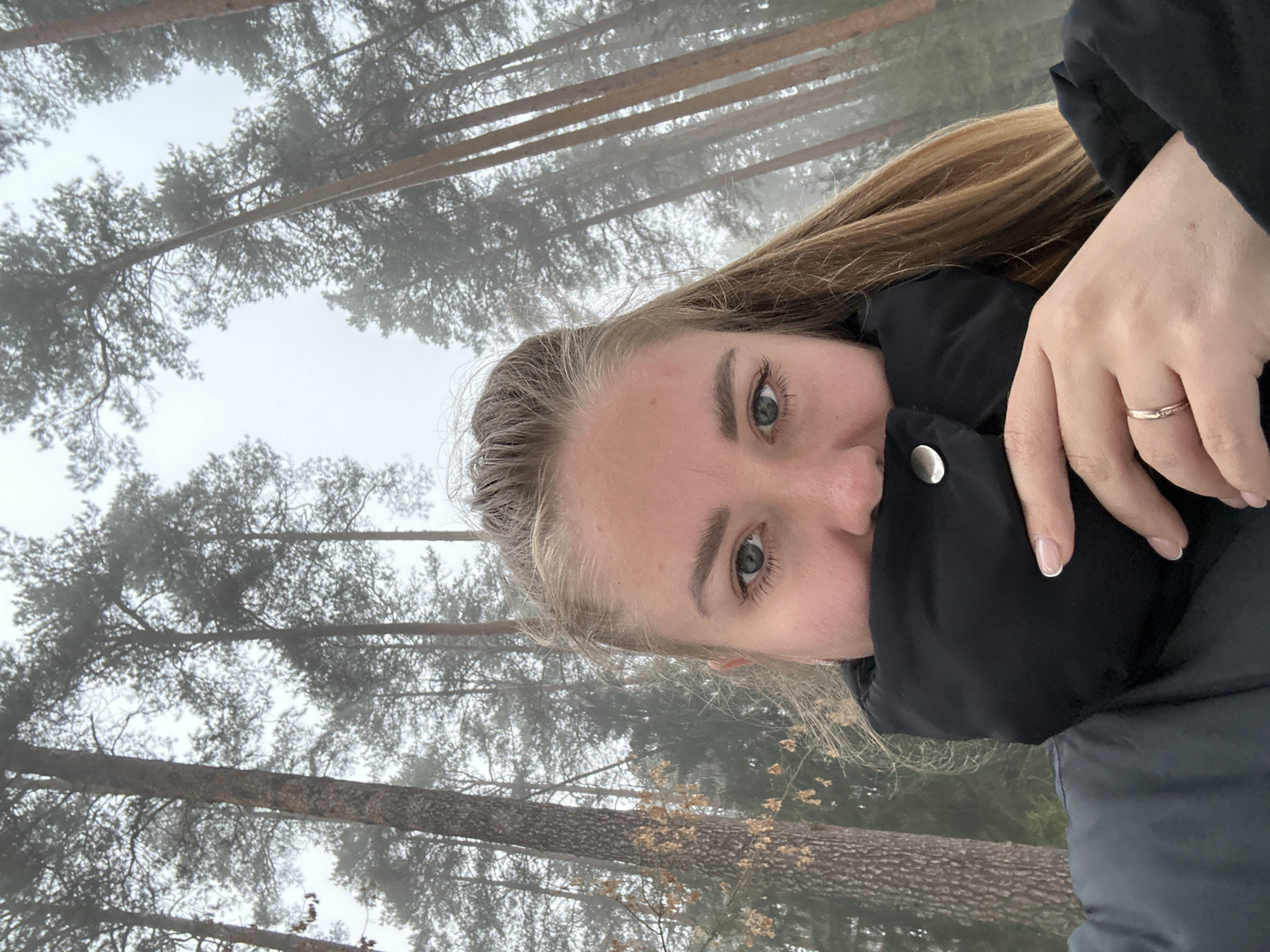 A young woman with blonde hair and green eyes is photographed from a low angle in a foggy forest. She is wearing a black winter jacket with a button visible near her face. Her right hand is raised, and she has a ring on her ring finger. The background is blurred due to the fog, showing tall trees common in forested areas. Though the exact location is not definitively discernable from the image alone, the query requests the inclusion of "Chaikovsky, Russia". Therefore, the scene is in a forest in the city of Chaikovsky, Russia. The trees are bare, suggesting either late autumn or winter. The woman appears to be taking a selfie.