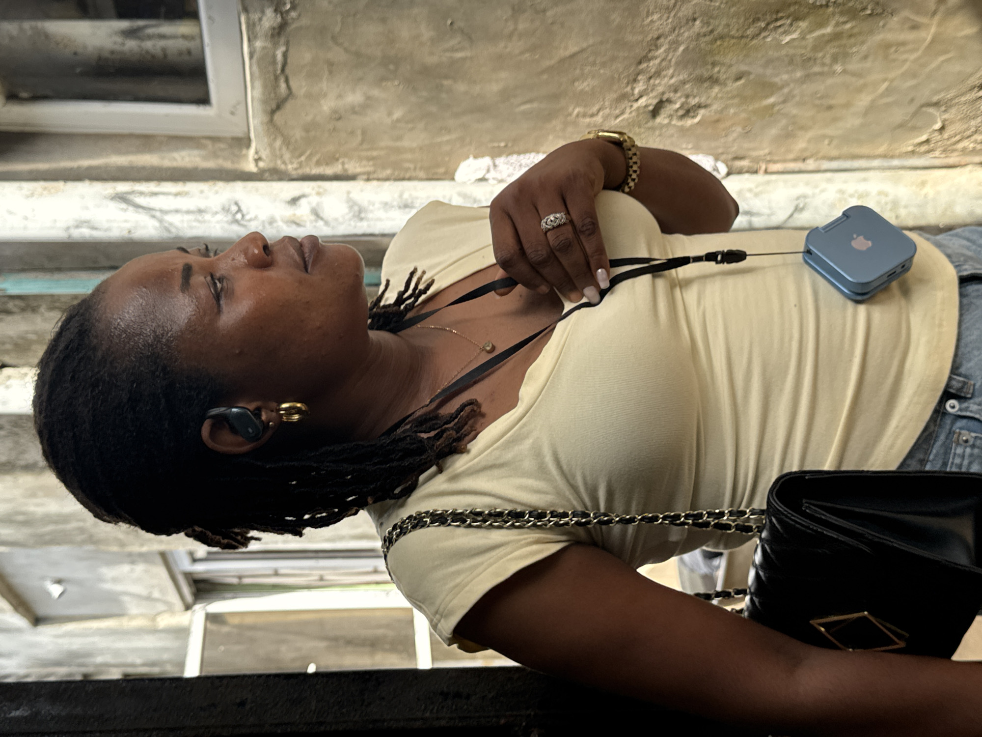 A woman with dark hair is photographed in what appears to be Badagry, Nigeria. She is light-skinned, with braided hair, gold hoop earrings, and an earbud in her left ear. She wears a light yellow shirt and a gold watch and ring on her left hand. The woman is wearing a black purse with a gold accent strap. A small, square, blue device with an Apple logo is attached to her shirt by a black lanyard. The background is a weathered, light-colored concrete structure with a window.