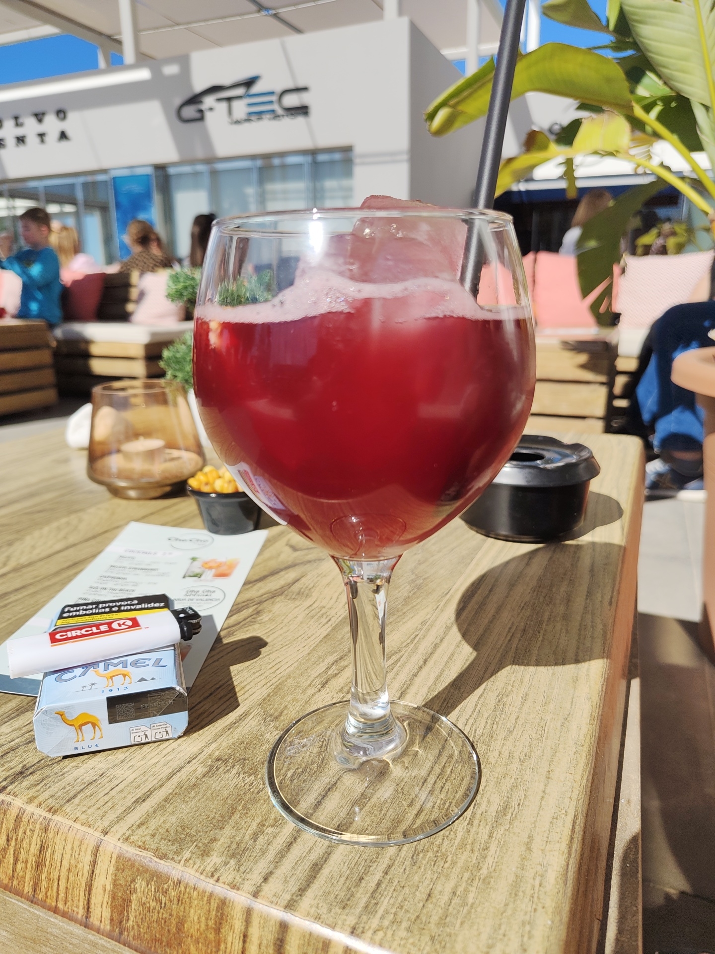 The image shows a close-up of a large wine glass filled with a red beverage and ice, likely sangria, sitting on a wooden table. A black straw protrudes from the glass. Next to the glass, on the left, is a pack of Camel Blue cigarettes with a white lighter on top, along with a menu card. There is a small black ashtray to the right of the glass.
In the blurred background, people are sitting at tables. A building is visible in the far background with "SLVO" and "ENTA" visible, as well as the logo "G-TEC." Lush green foliage is to the right of the glass.
The scene appears to be at an outdoor cafe or restaurant during the day in Dénia, Spain.