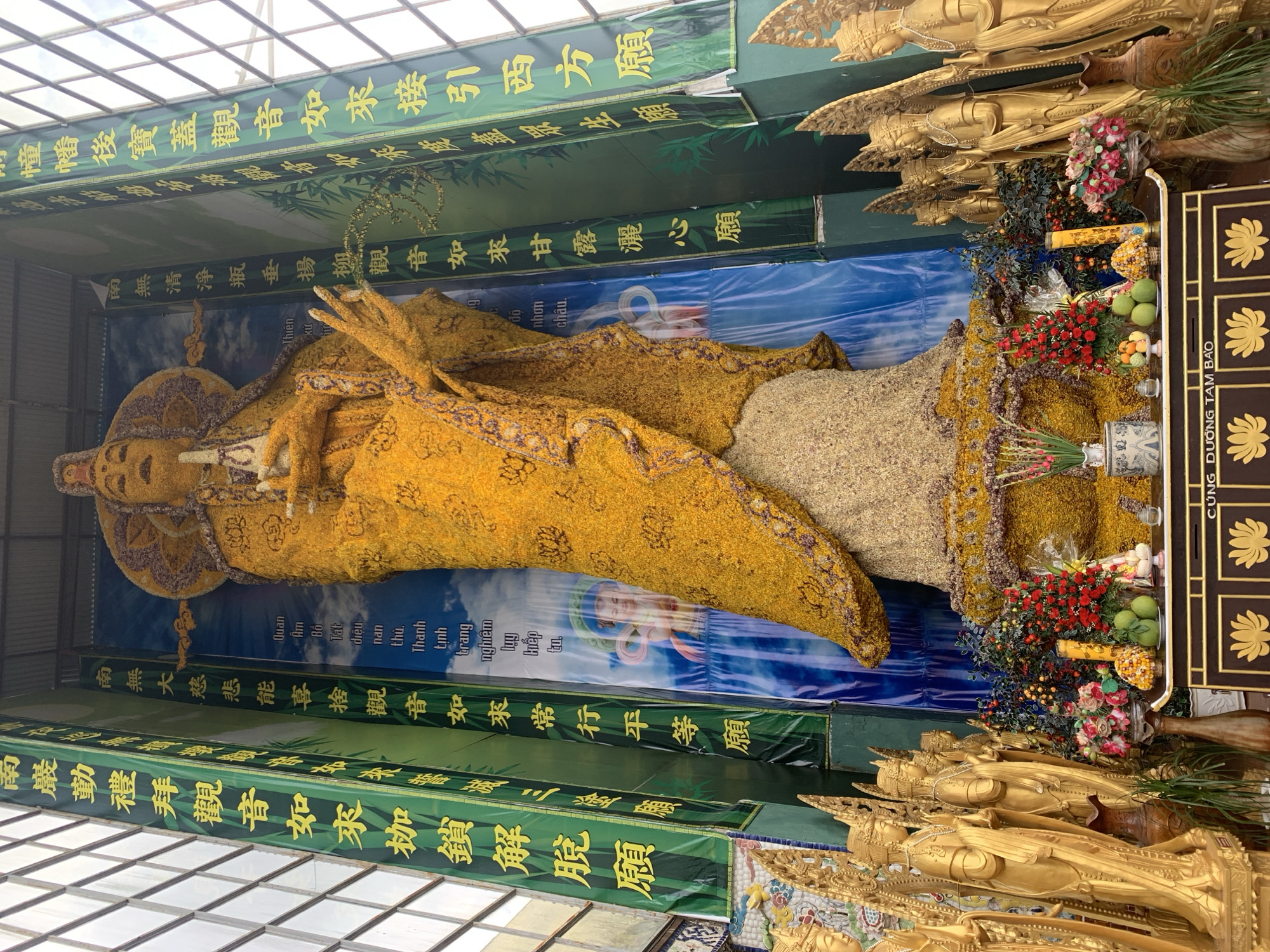The image shows a large, yellow, flower-covered statue of a deity, likely Guan Yin, in Xuân Thành, Vietnam. The statue is adorned with many small yellow flowers and is situated in a temple-like setting. Flanking the statue are tall green banners with yellow characters, possibly religious inscriptions. To the left and right of the main statue, there are rows of smaller, golden statues of deities or attendants. In the foreground, a dark, ornate altar is laden with offerings of fruit, flowers, and candles. Behind the main statue, a blue backdrop with clouds and images of children is visible. The entire scene is lit by natural light, suggesting an indoor or partially covered space.