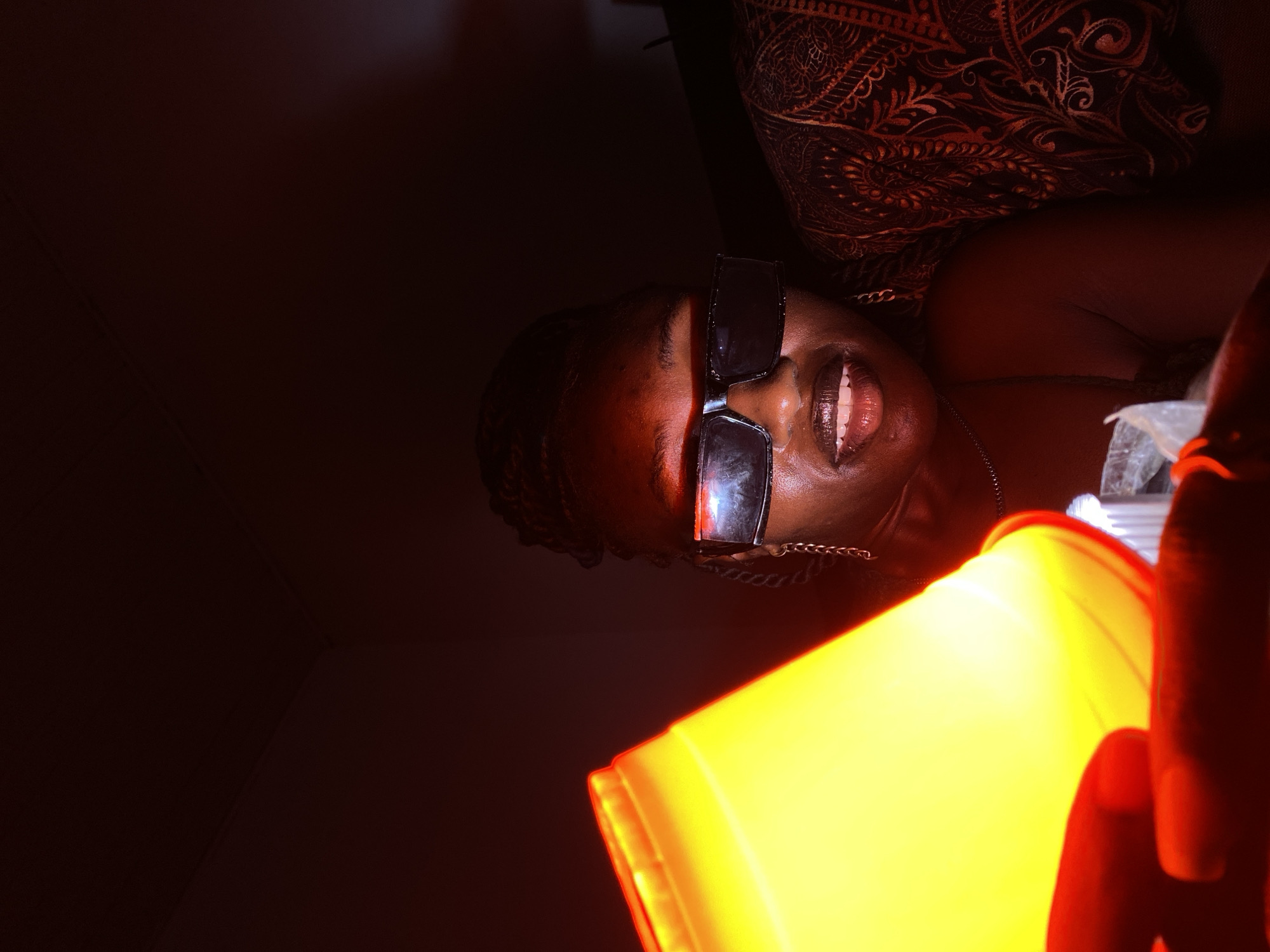 The image shows a young woman wearing dark rectangular sunglasses and a silver necklace. She has dark braided hair and is smiling, revealing her teeth. She is holding a bright yellow object, possibly a light source or a decorative item, in her left hand, which casts a strong yellow glow on her face and the surrounding area. The background is dark, with a hint of a patterned fabric visible on the right side. The lighting suggests an indoor setting, possibly at night. The woman's facial expression and the way she is holding the object indicate she is posing for the photo. There is no discernible information within the image to suggest the specific location in the city of Bar Jirgi Summa, Nigeria.