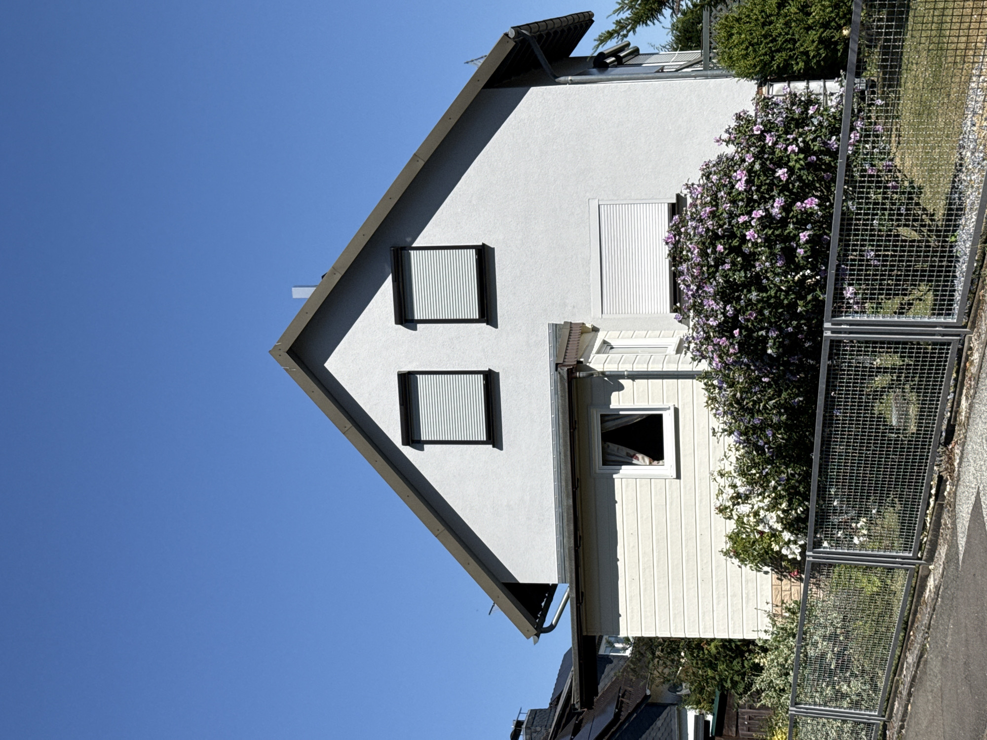 The image depicts a two-story house with a light gray stucco exterior and a dark brown roof. Two windows with white roller shutters are visible on the upper level, and another larger window with roller shutters is on the lower level. A section of the house on the left has a light beige, horizontal siding. A metal fence runs along the front of the property, with a dense bush of purple and white flowers in front of it. The sky is clear and blue. This house is located in the city of Bad Sachsa, Germany.
