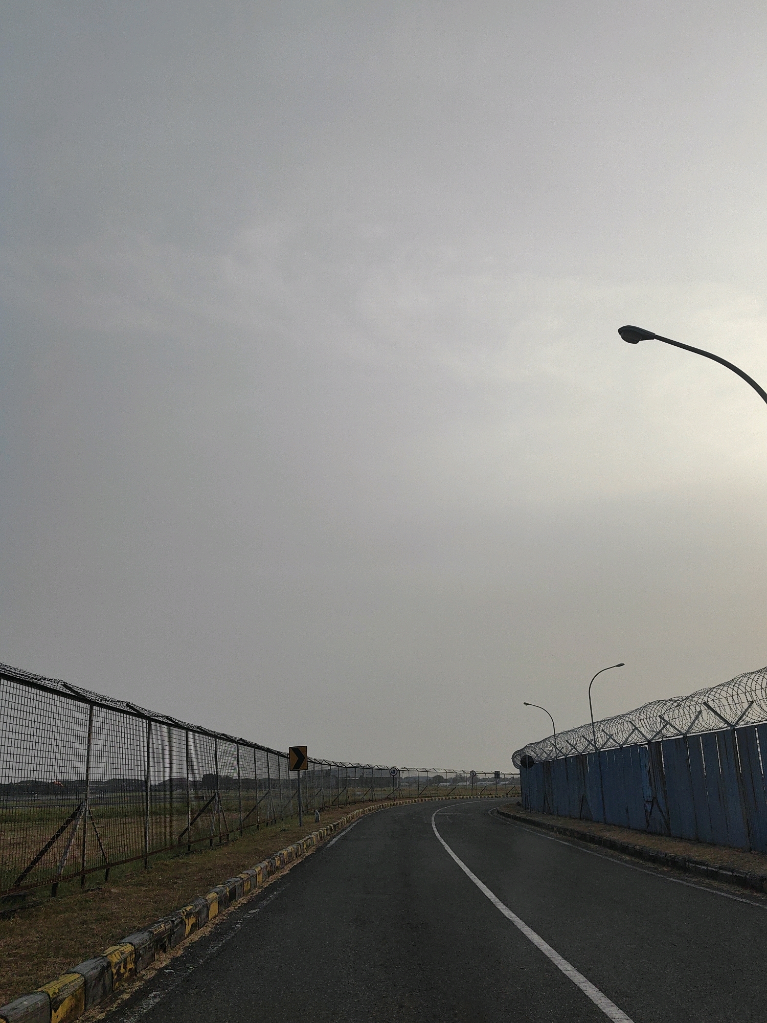 The image depicts a road curving through an area bordered by high fences topped with barbed wire. On the left, a chain-link fence runs along the road's edge, backed by dry grass and some vegetation. A yellow and black striped curb runs along the road. On the right, a solid blue fence, also topped with barbed wire, borders the road. Two streetlights are visible, one in the upper right and another further down the road on the right. The sky is overcast and pale. The road itself is asphalt with a white line marking the center. In the distance on the left, faint structures and what appear to be aircraft can be seen. The scene suggests an airport perimeter or a secure industrial zone. The location is identified as Betro, Indonesia.