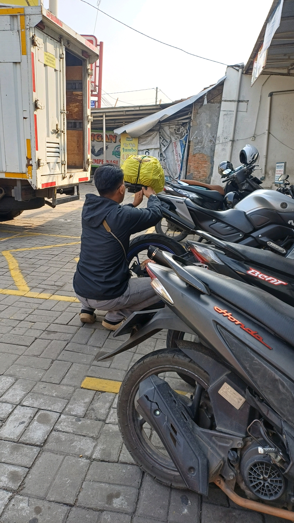 In Gunung Putri, Indonesia, a person wearing a black hoodie and grey pants squats down to secure a bright yellow bundle onto the rear rack of a parked motorcycle. The person is facing away from the camera, their back towards a white truck with an open cargo door on the left side of the frame. Behind the person, several motorcycles are parked in a row, their seats visible. The ground is paved with interlocking gray bricks, marked with yellow lines. In the background, a low-slung building with a corrugated metal roof and partial walls stands under a bright sky. The scene appears to be an outdoor parking area, possibly a commercial or transportation hub.