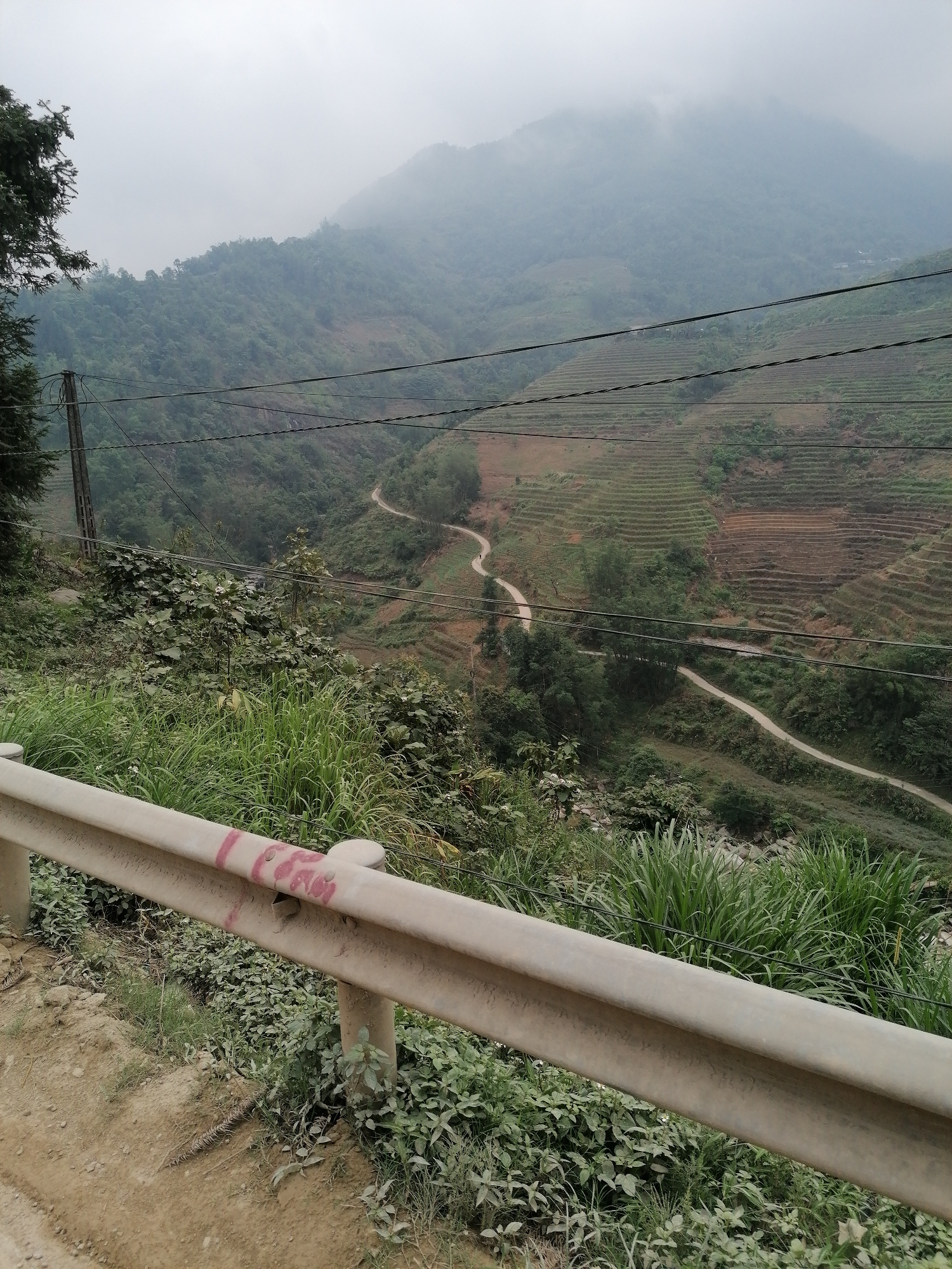 The image shows a mountainous landscape on a cloudy day, likely in a rural area of Unknown. A white guardrail is visible in the foreground, with green foliage growing around it. In the background, a winding dirt road traverses a terraced hillside. Power lines stretch across the scene, and a mountain is partially obscured by fog. No people are visible. The setting appears natural and undeveloped.