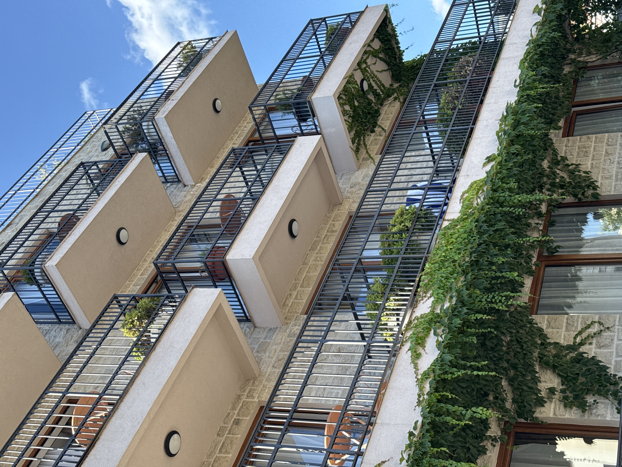 The image shows a close-up, low-angle view of a modern building facade with a striking architectural design. The building features a series of staggered balconies, each enclosed by dark metal railings and shaded by angled, light-colored overhangs. These overhangs create a geometric pattern that repeats up the side of the structure.
Lush green ivy is climbing the building, cascading down the white stone walls and over some of the balconies, adding a natural element to the contemporary architecture. The windows are visible behind the metal railings, some with reflective surfaces showing glimpses of the sky and surrounding environment.
The sky above is blue with scattered white clouds, suggesting it is daytime, likely late morning or early afternoon due to the bright lighting and clear visibility. The overall scene conveys a sense of modern, urban living with an emphasis on design and integration with nature. The location cues, such as the specific architectural style and the presence of the ivy, are consistent with coastal European architecture. No people or specific activities are depicted. There is no visible text in the image.